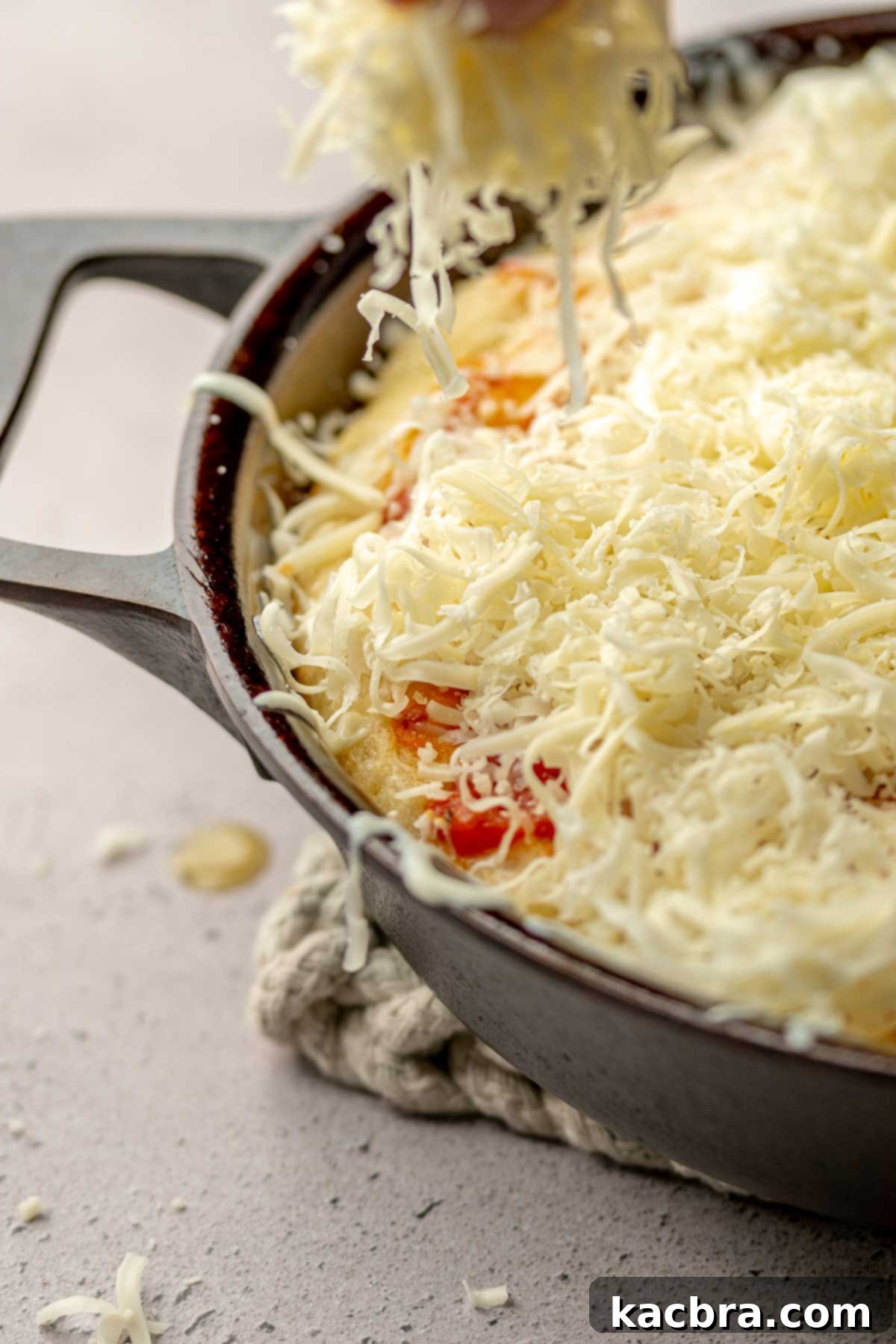 A hand adds shredded cheese on top of the pizza sauce.