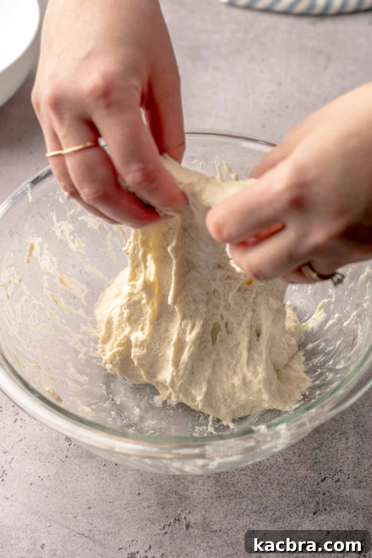 Two hands stretch dough upwards in a bowl.