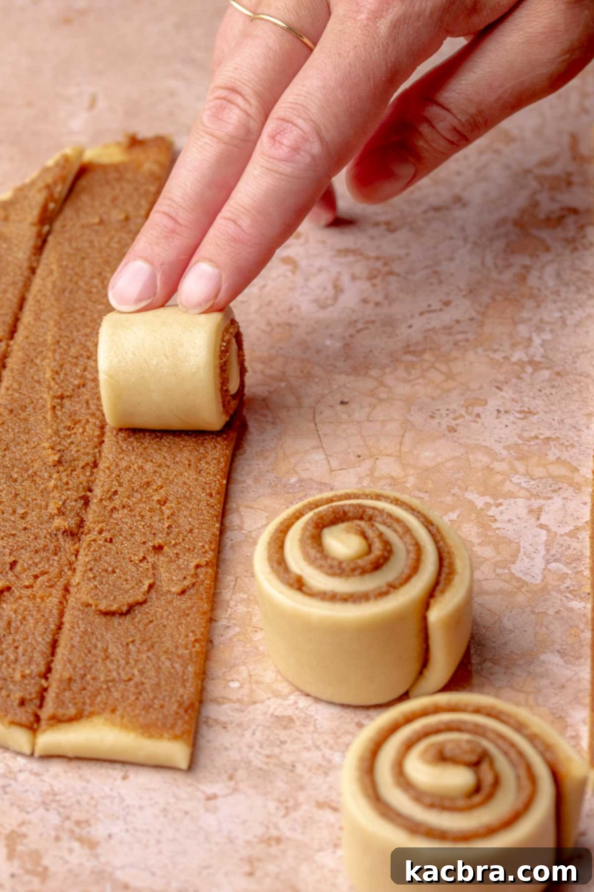 A hand rolls up strips of cinnamon roll dough into small coils.