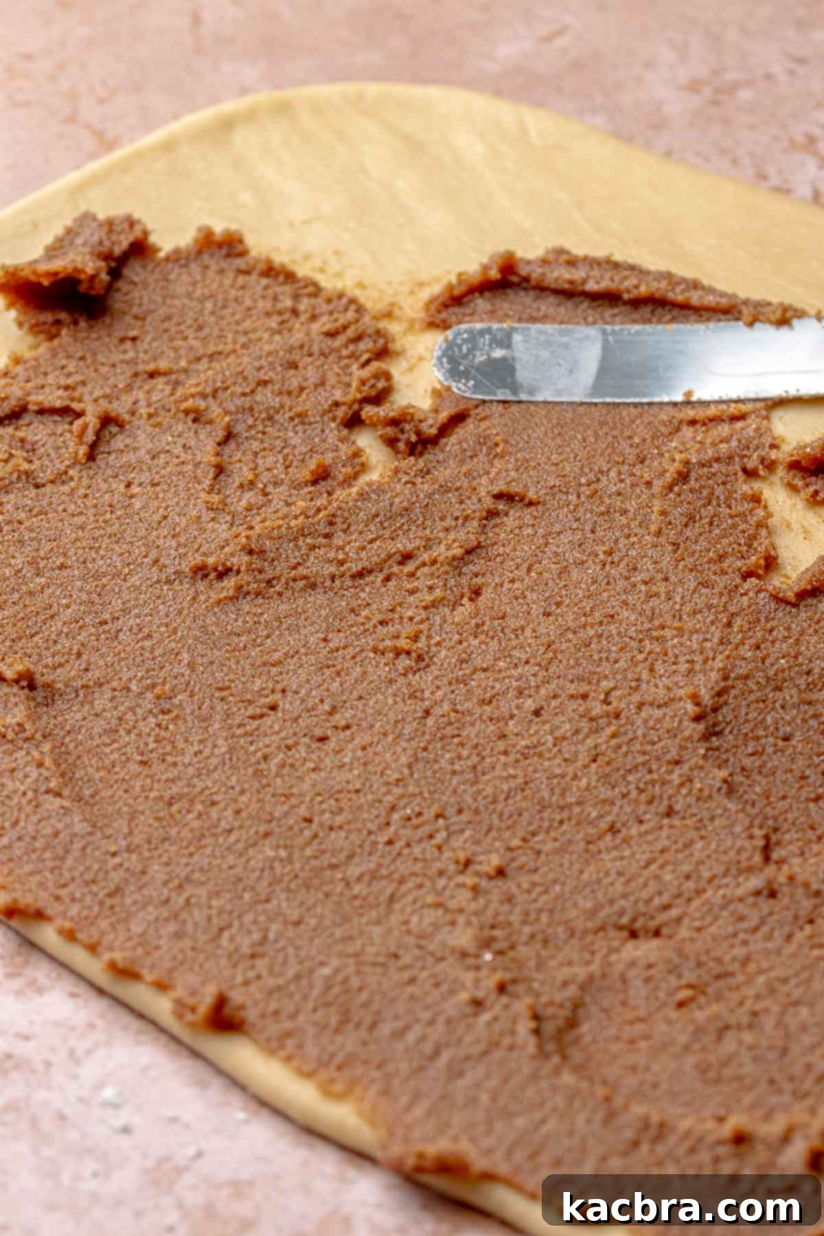 An offset spatula spreads cinnamon sugar filling onto a cinnamon roll dough.