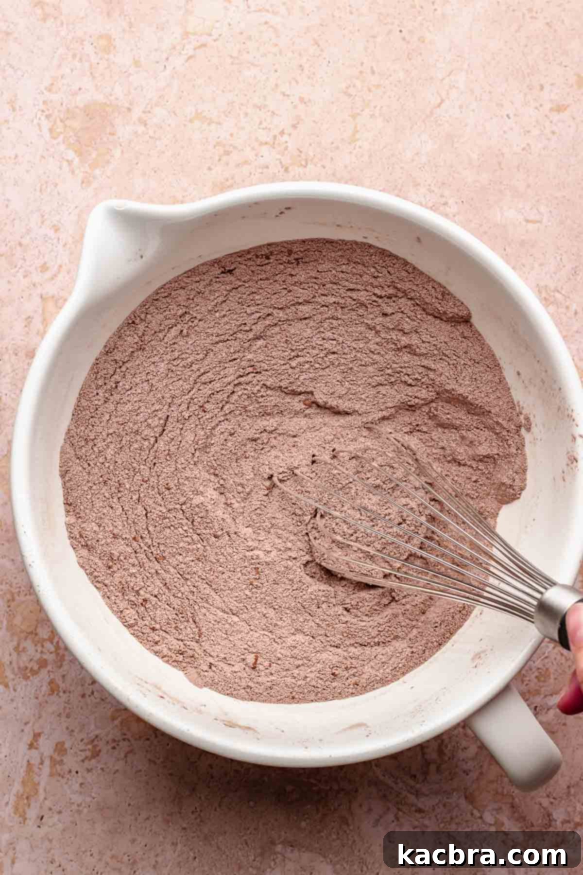 A whisk mixes dry chocolate cake ingredients in a bowl.