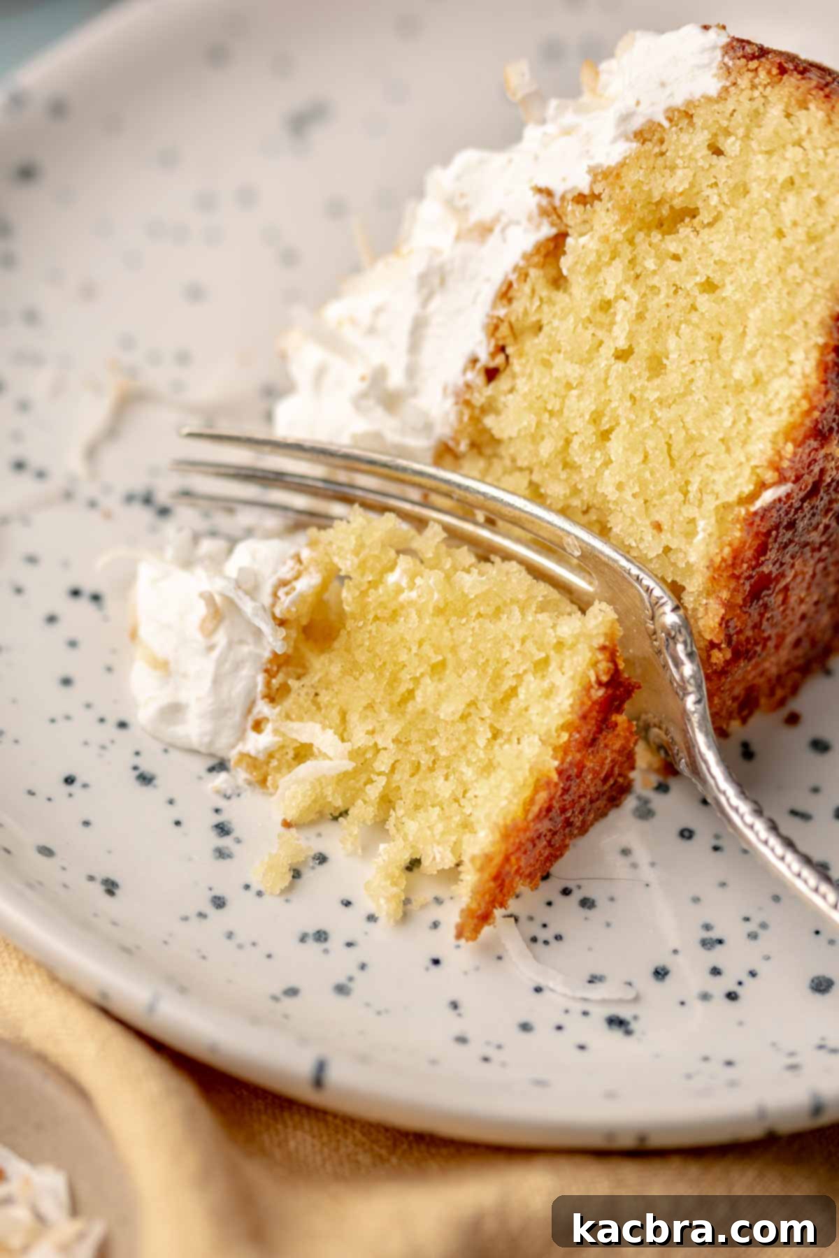 A fork cuts through a piece of coconut almond cake on a plate, revealing its moist interior.