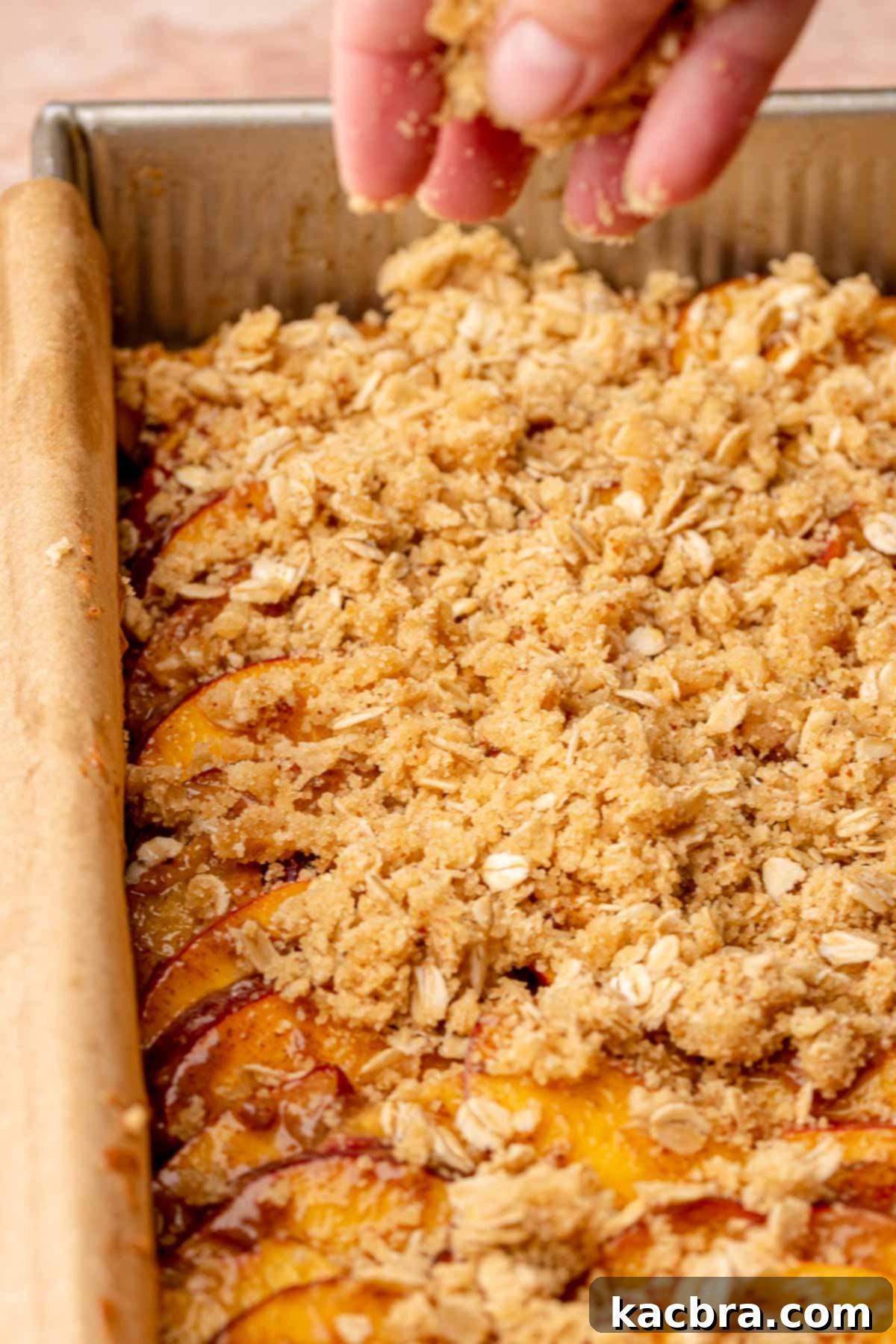 A hand adding the final layer of oatmeal crumble onto peach bars, preparing them for baking.