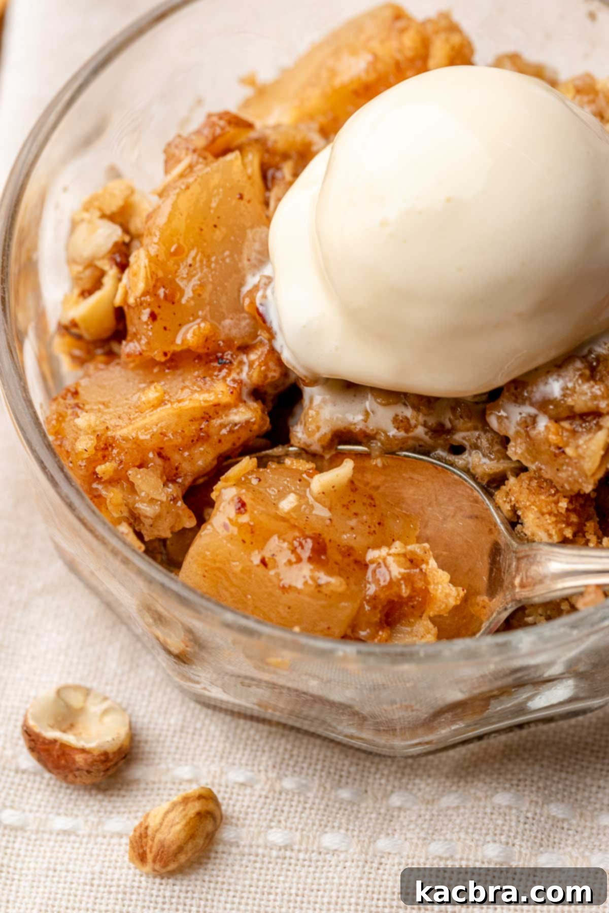 Warm Toffee Apple Hazelnut Crumble in a bowl topped with melting vanilla ice cream and a spoon.