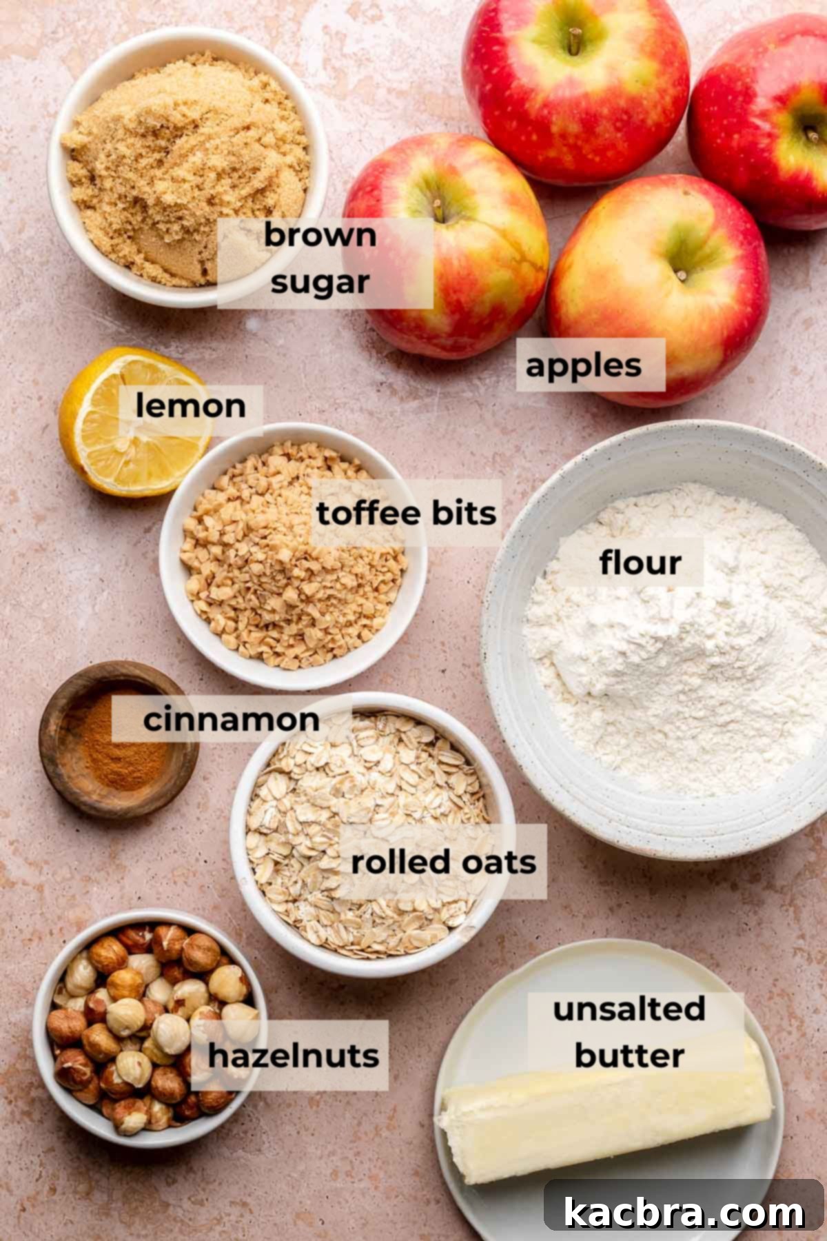 Various ingredients for Toffee Apple Hazelnut Crumble laid out on a table, including apples, oats, butter, sugar, and hazelnuts.