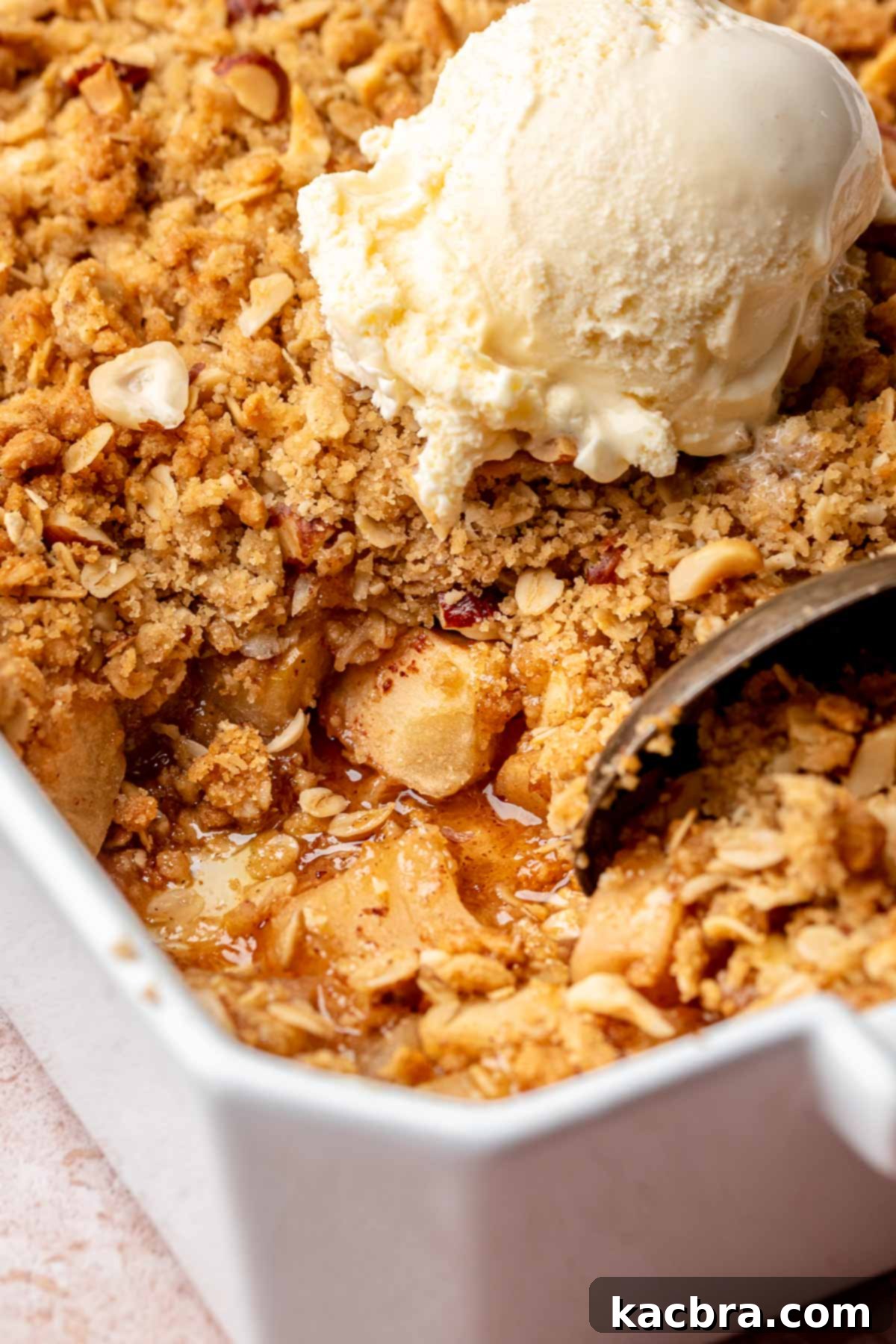 A spoon scoops a portion of Toffee Apple Hazelnut Crumble from a large casserole dish, showing the gooey filling and crunchy topping.