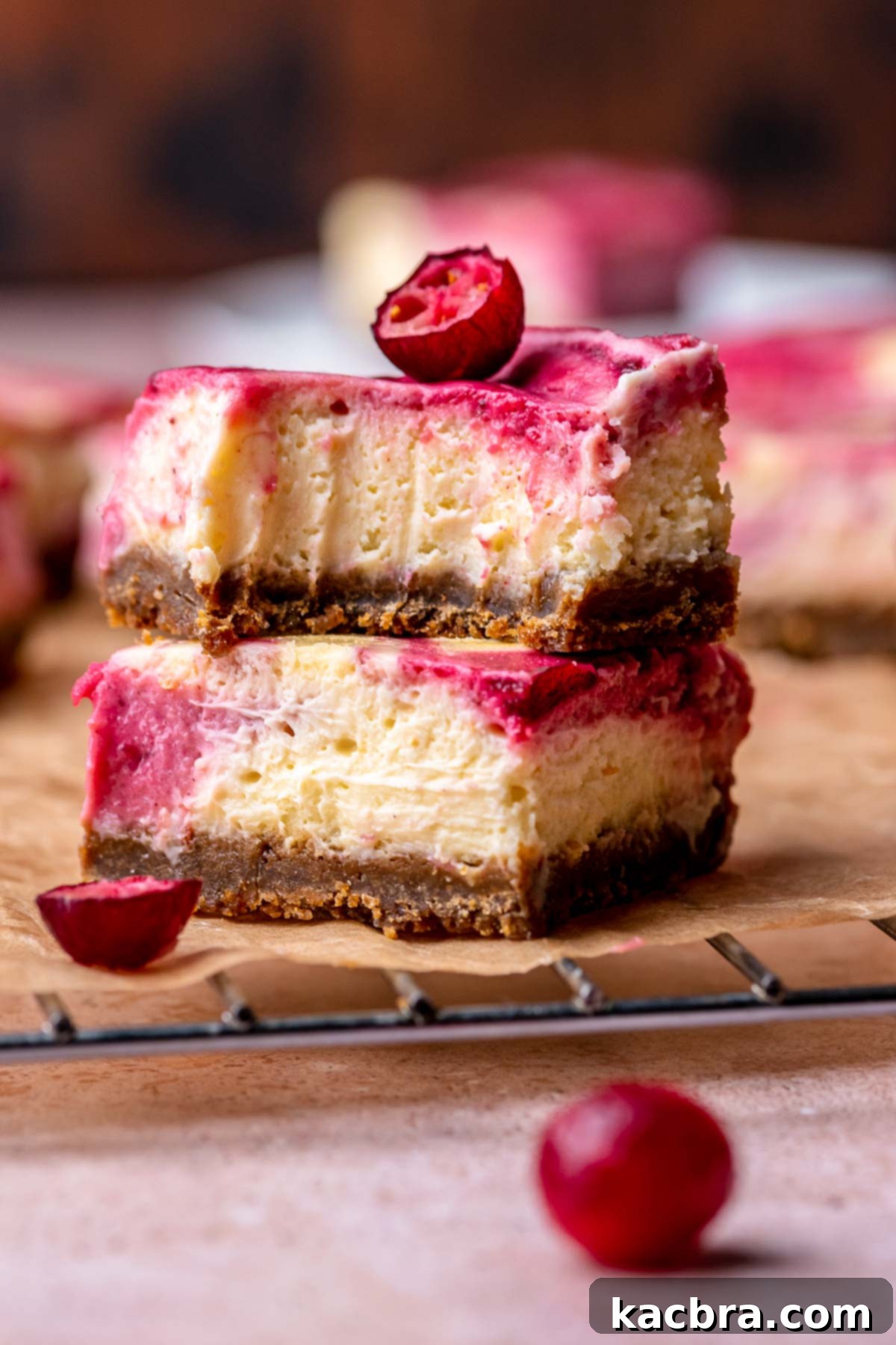 Two vibrant cranberry cheesecake bars, stacked on a piece of parchment paper, with a bite taken out of the top bar to reveal the creamy texture and swirled layers.