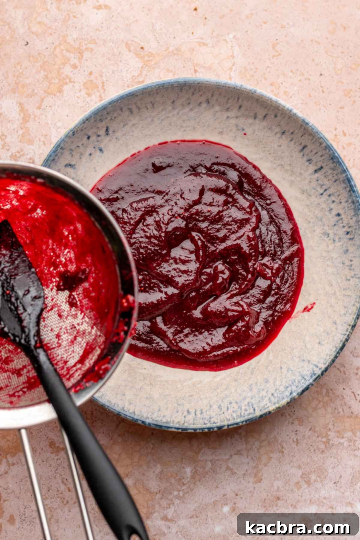 A spatula gently pushes the vibrant cranberry puree through a fine mesh sieve into a clean bowl, ensuring a smooth, seedless sauce.