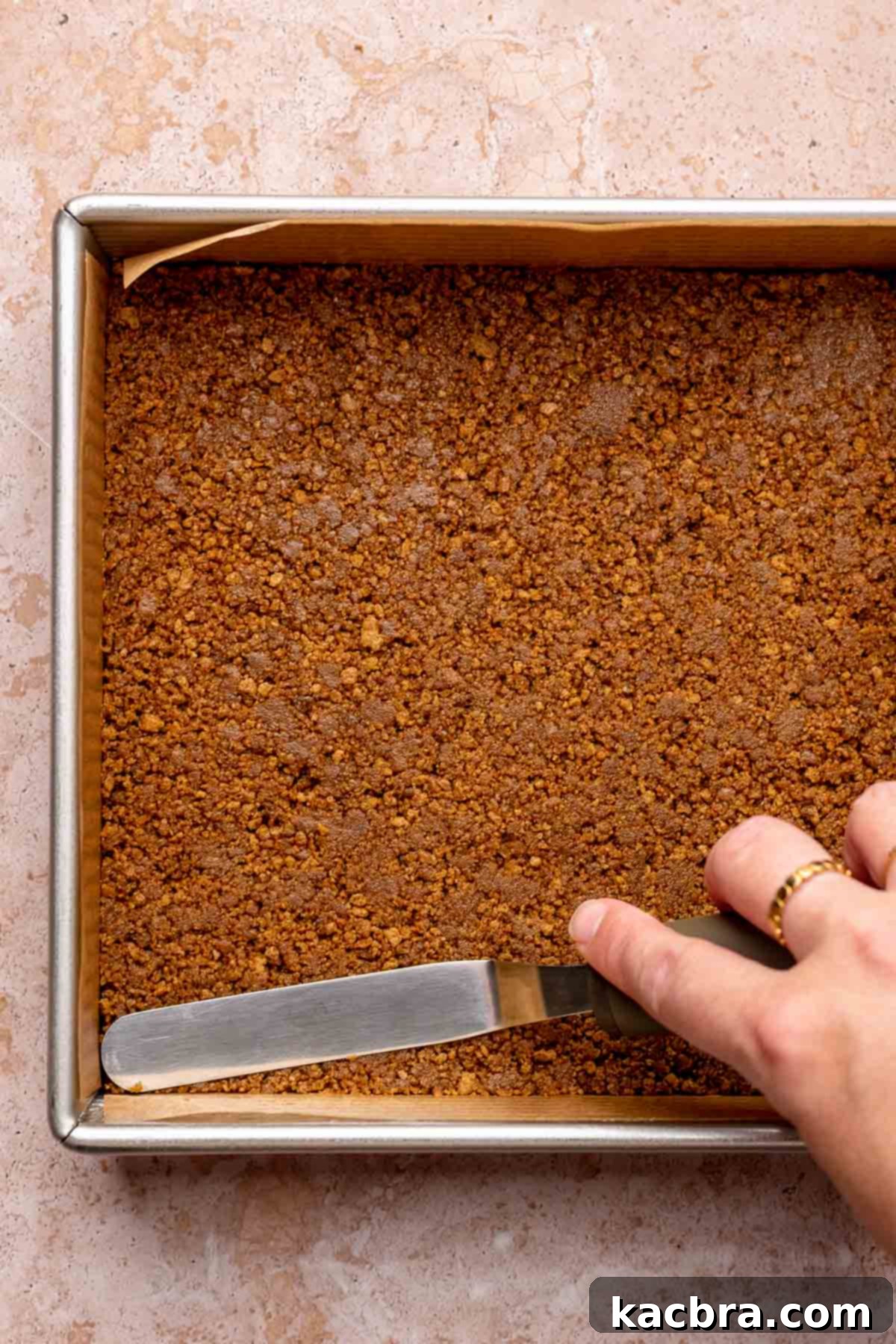 An offset spatula evenly presses the spiced gingersnap crust into a square baking pan, forming a compact base for the cheesecake.