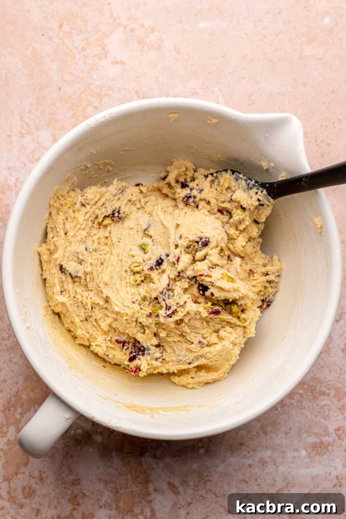 A spatula gently folding chopped pistachios and dried cranberries into the biscotti dough, ensuring even distribution.
