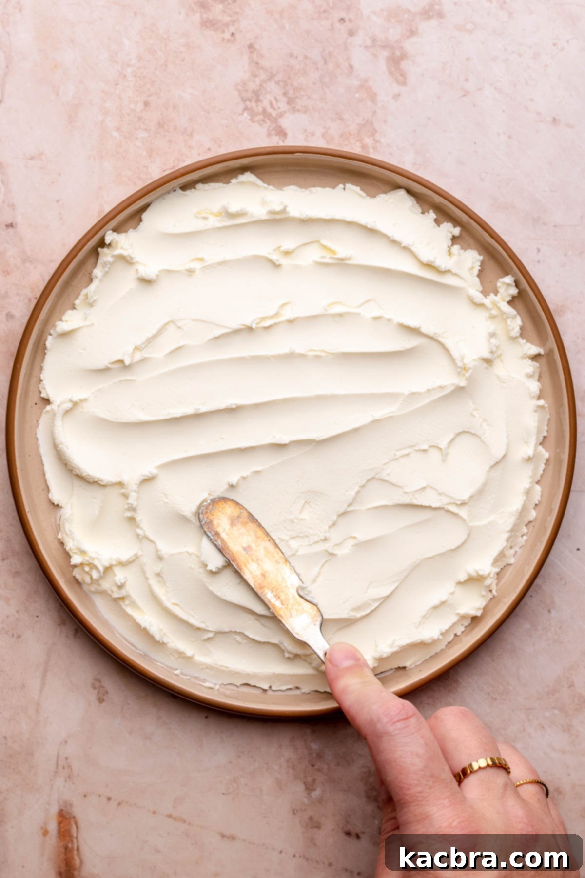 A knife spreads softened cream cheese evenly onto a round serving plate.