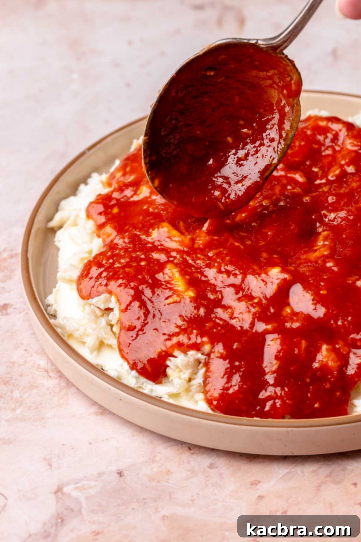 A spoon drizzles the prepared cocktail sauce over the crab and cream cheese layers.