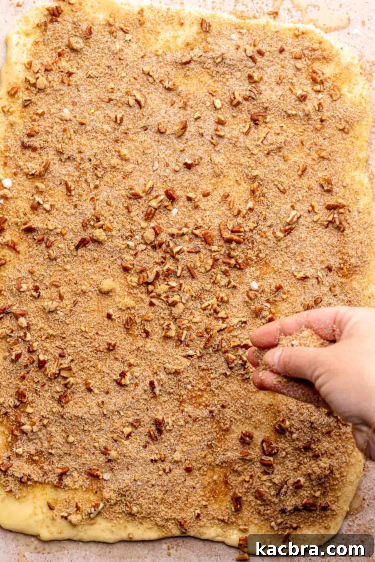 A hand sprinkling cinnamon sugar and chopped pecans onto buttered dough.