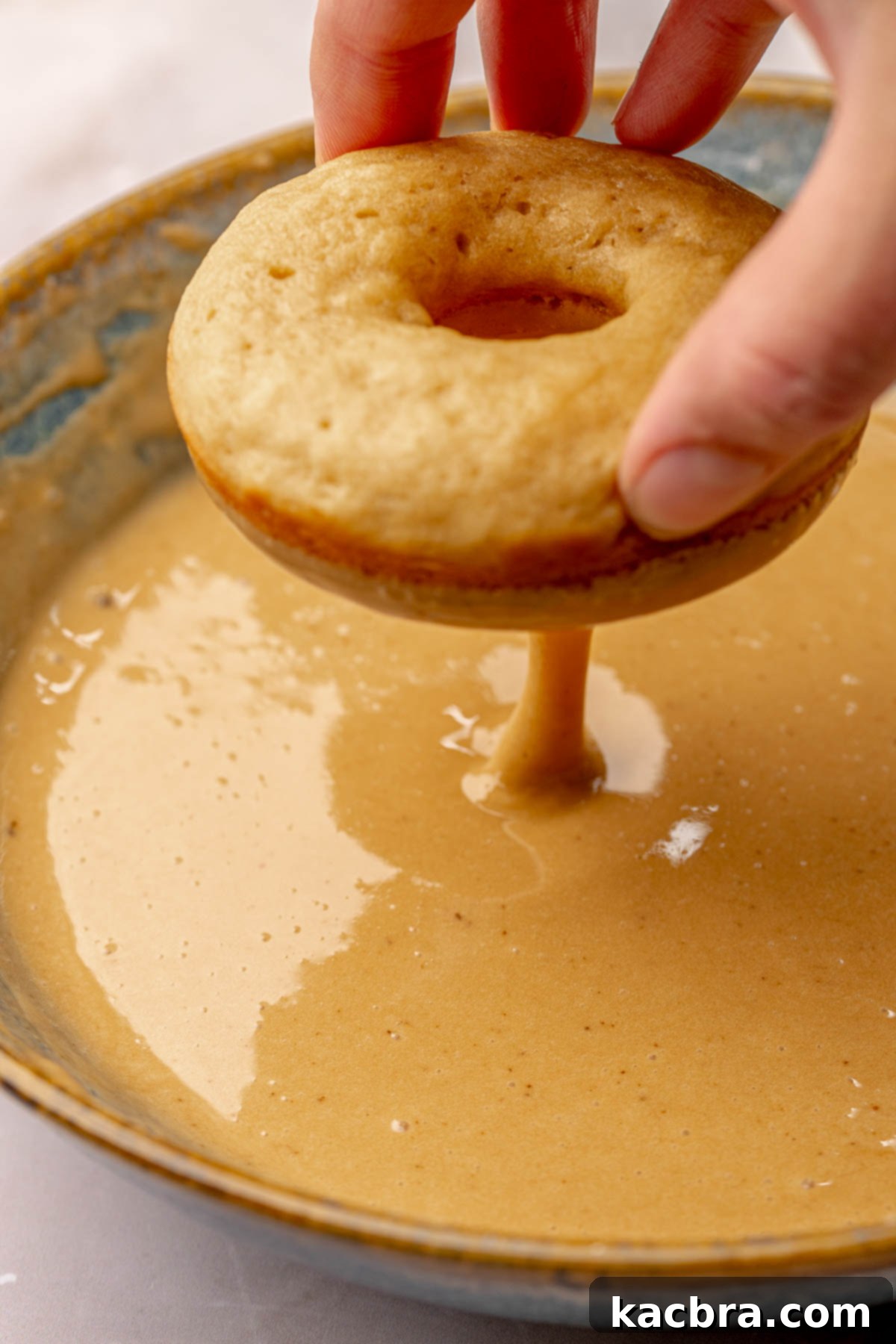 A donut is dipped in coffee glaze and the glaze is dripping off, ready for toppings.