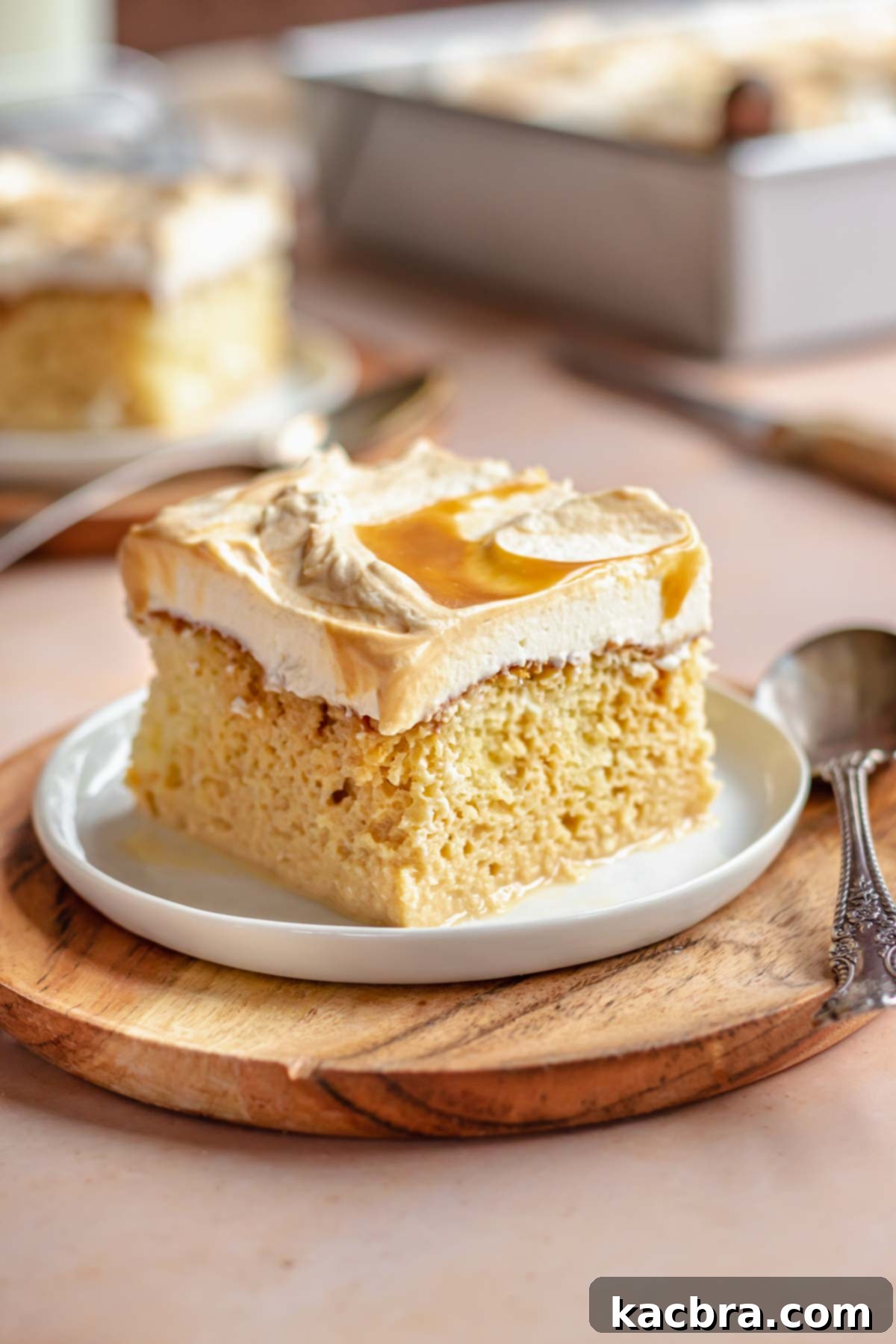 A slice of caramel tres leches cake on a white plate, garnished with a spoon and a hint of dulce de leche drizzle.