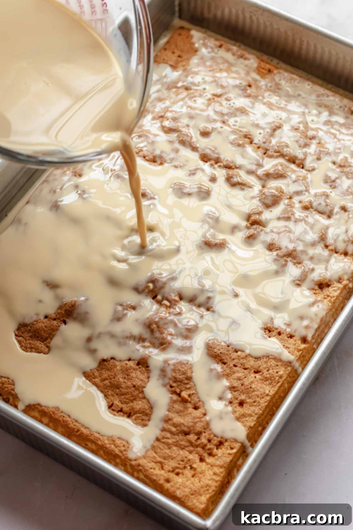 The rich caramel-infused milk mixture being poured evenly over the porous, slightly cooled baked cake in a pan.