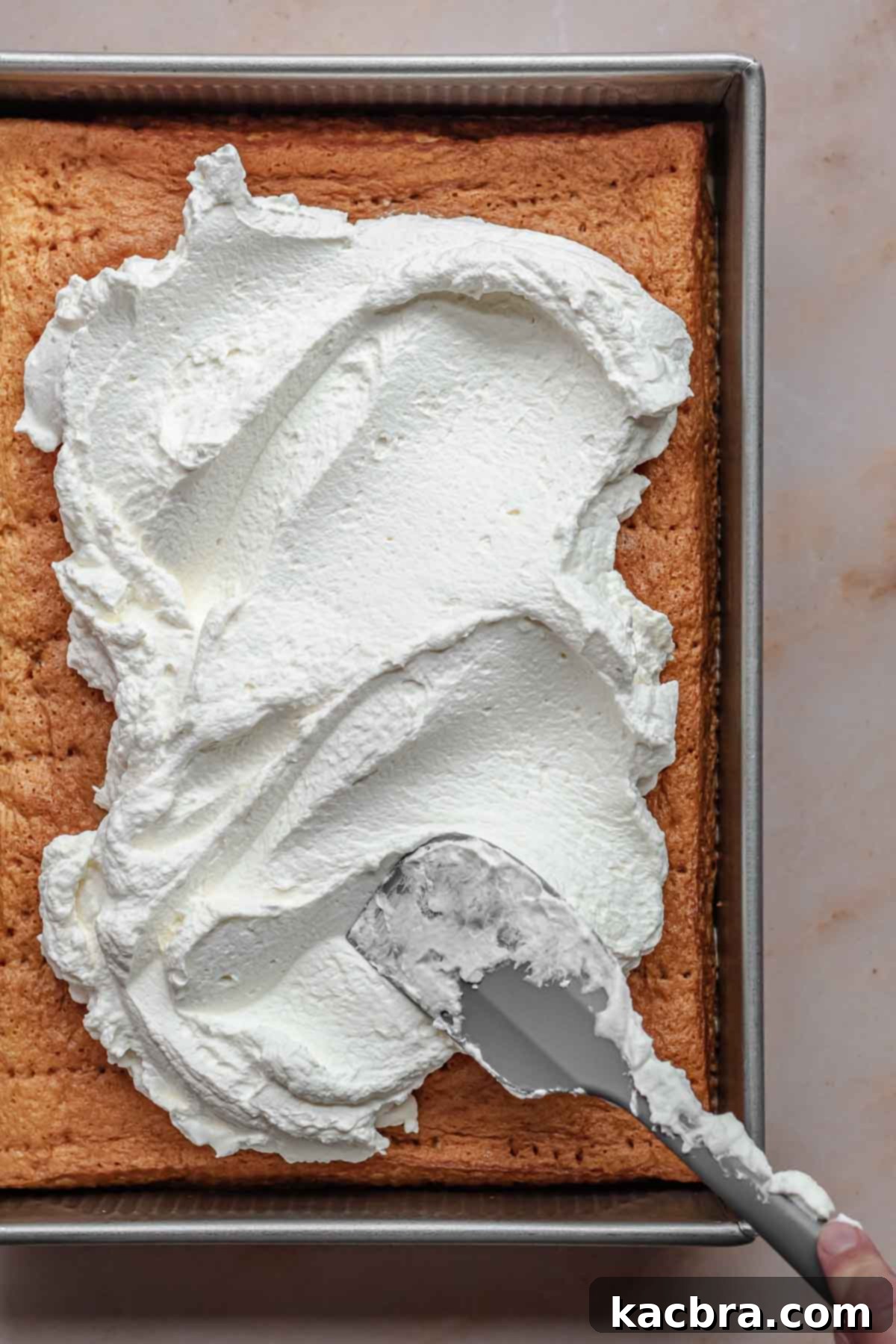 A spatula elegantly spreads a thick layer of freshly whipped cream over the surface of the soaked caramel tres leches cake.