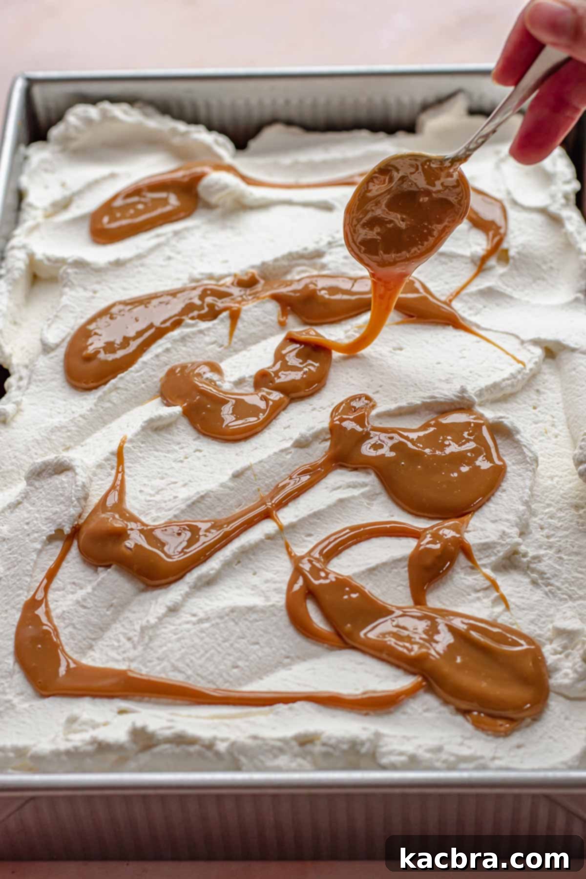 A spoon delicately drizzles the remaining thick dulce de leche over the fresh, white whipped cream topping of the cake.