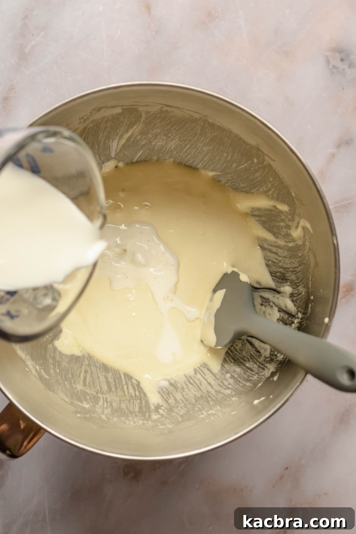 Whole milk being carefully poured into a mixing bowl containing pale yellow, fluffy beaten egg yolks and sugar.