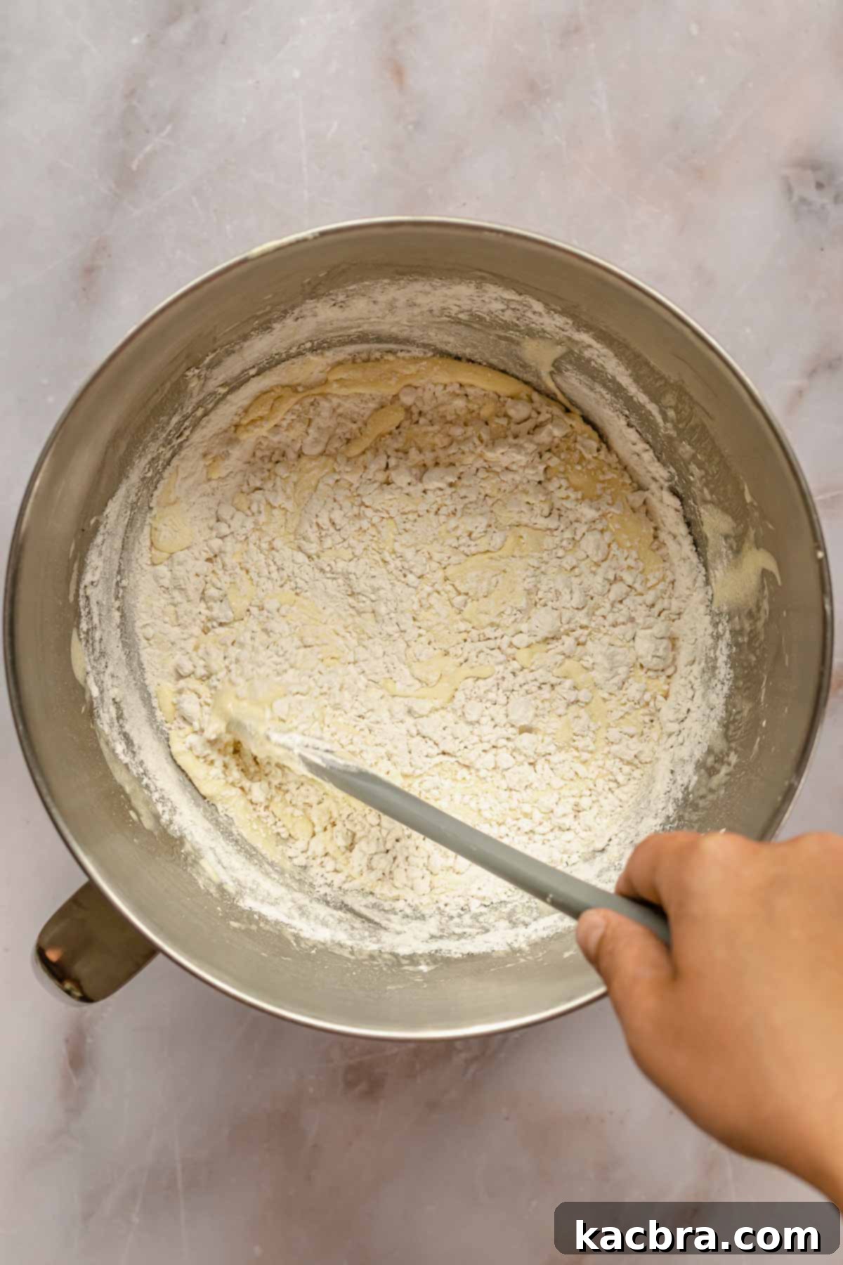 A spatula gently folds flour into a bowl of light yellow cake batter, demonstrating the proper folding technique.