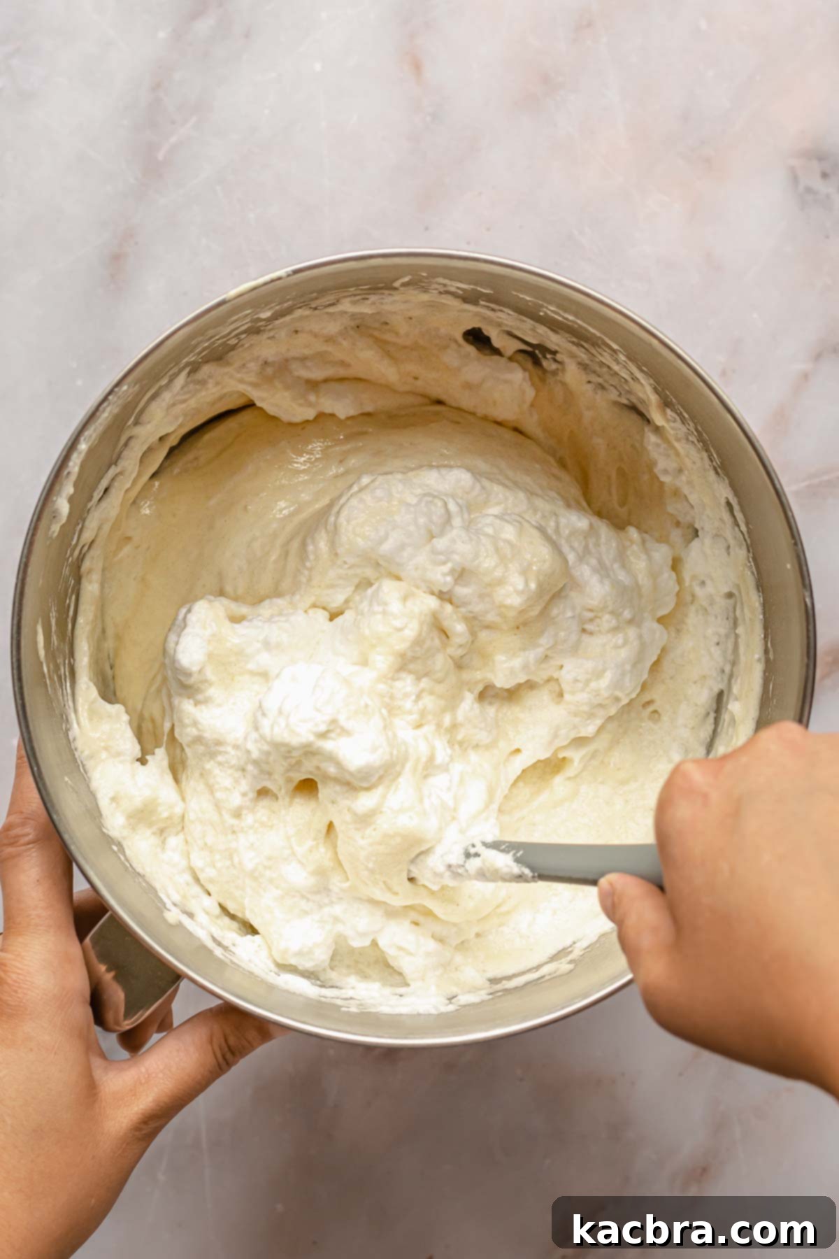 A hand gently folds whipped egg whites into a bowl of cake batter, incorporating air to keep the cake light.