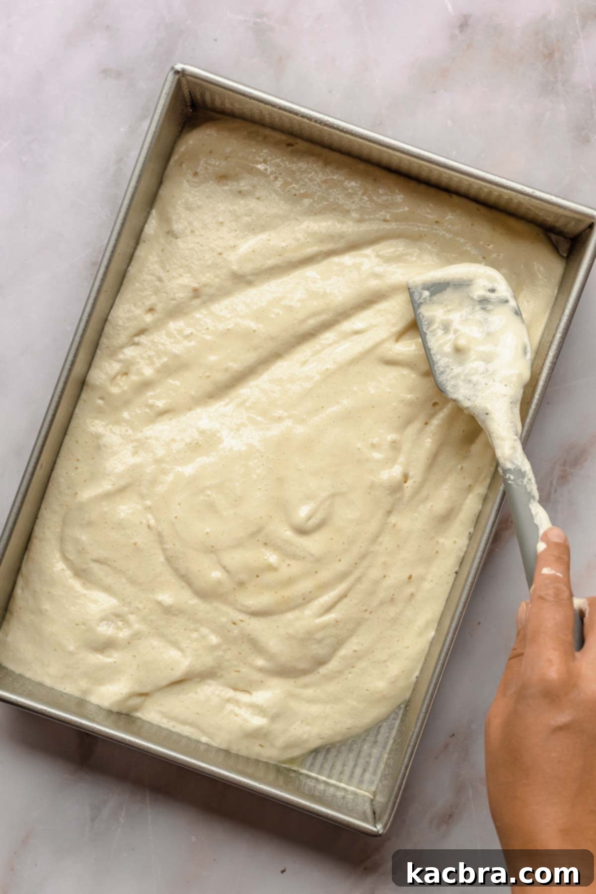 A spatula smoothly spreads light, airy cake batter into a large rectangular baking pan, ensuring an even layer.