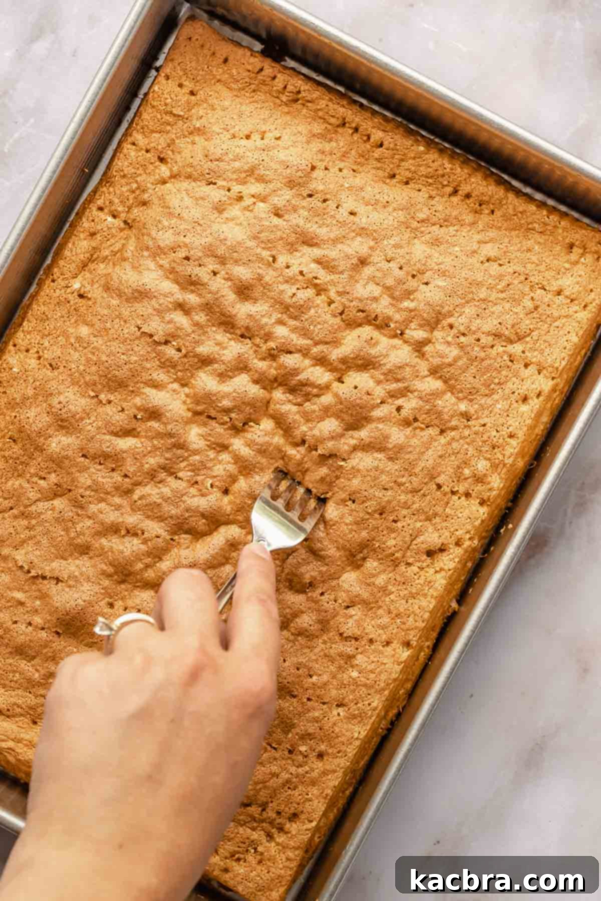 A fork creates numerous holes across the surface of a freshly baked sponge cake, preparing it for soaking.