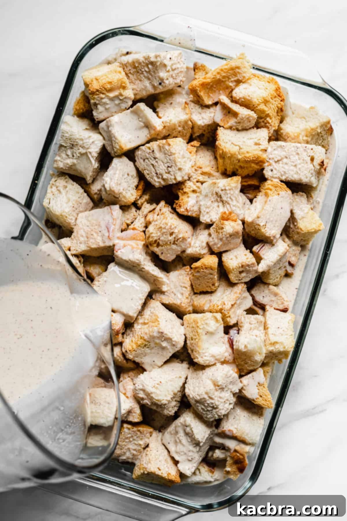 Rich custard being poured over toasted bread cubes and pecans in a casserole dish.