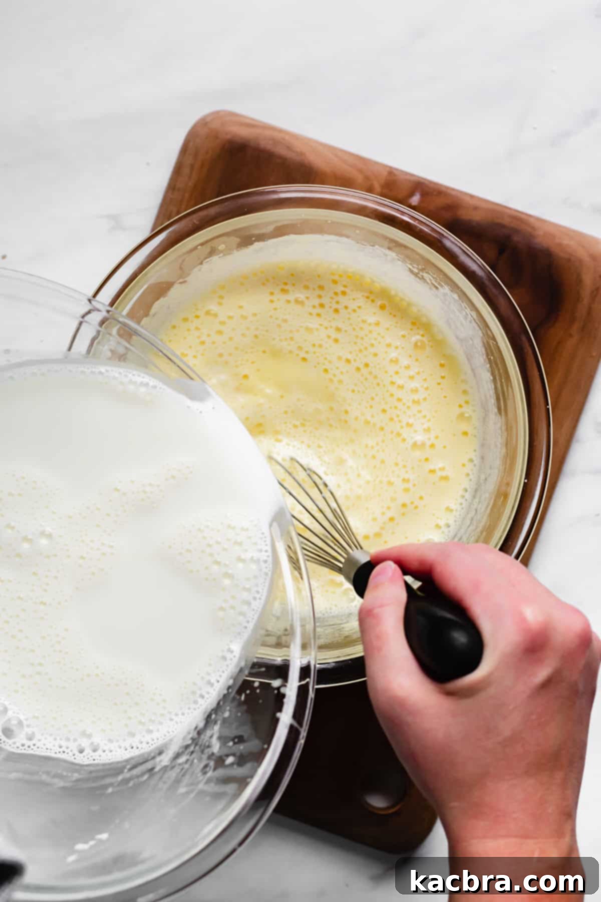 Hot milk being slowly poured into whisked egg yolks and sugar, demonstrating tempering technique.