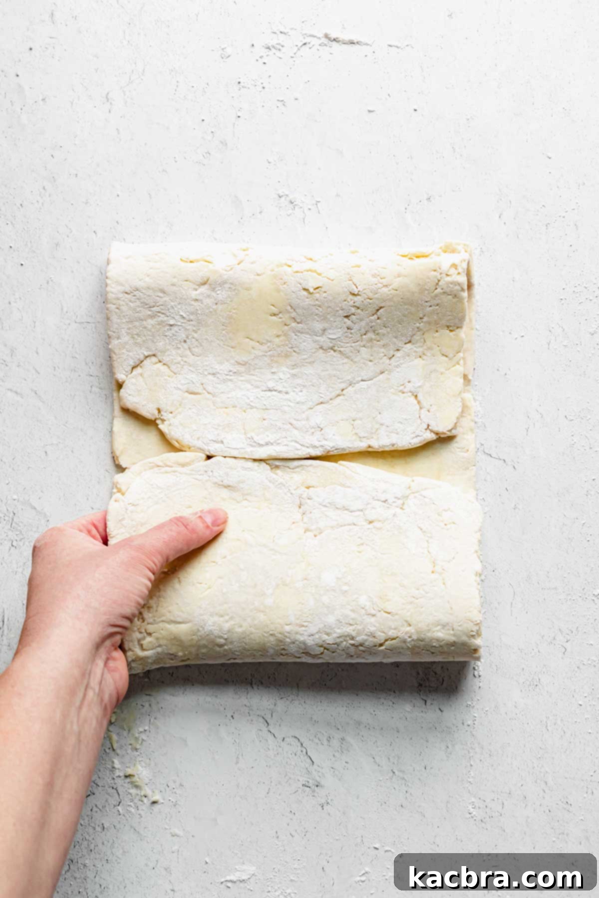 A hand folding the bottom quarter of the pastry dough up towards the center line, meeting the top fold.