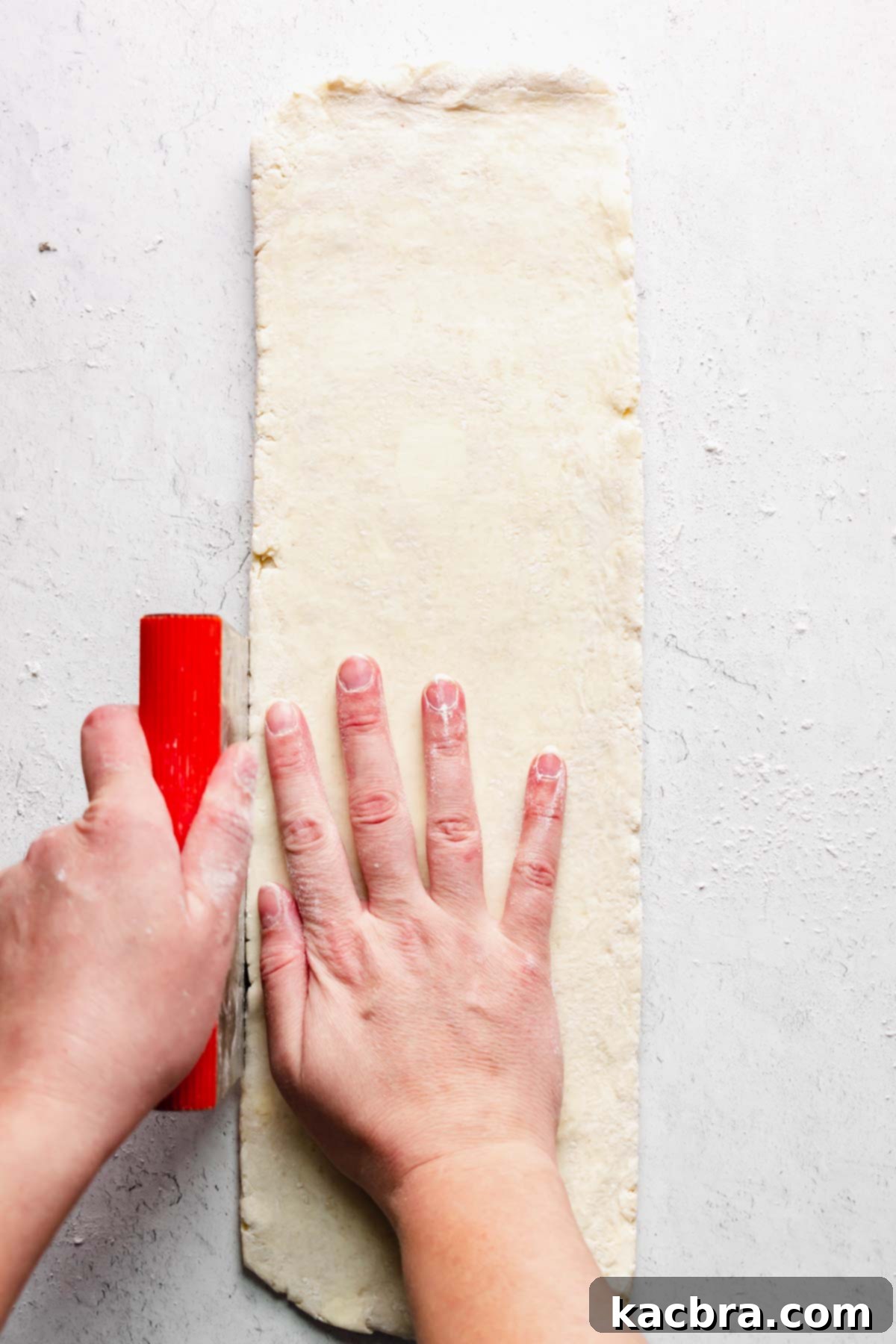 A long rectangle of dough with a hand and bench scraper cleaning up the sides.