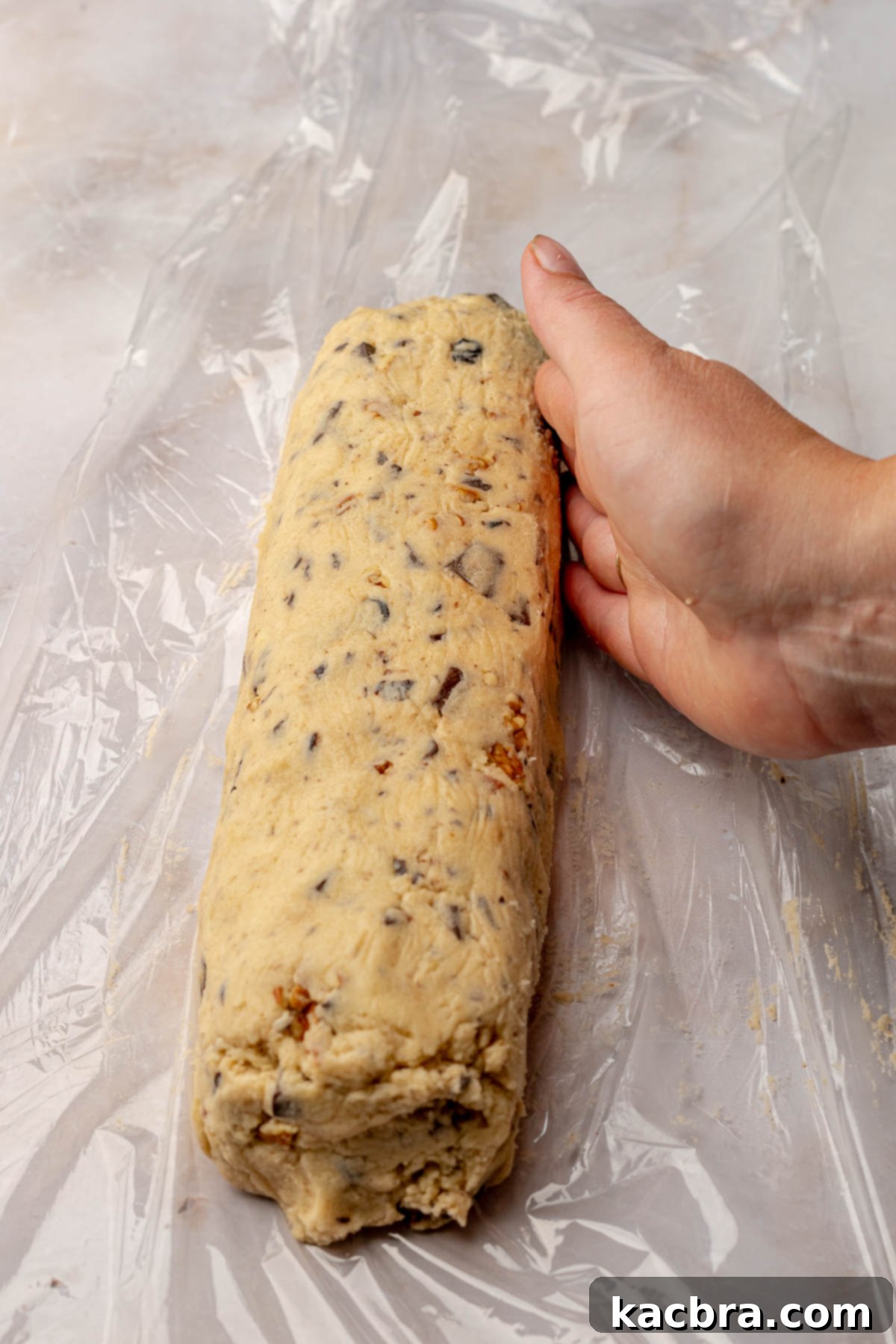 A hand forms cookie dough into a uniform log on a piece of plastic wrap.