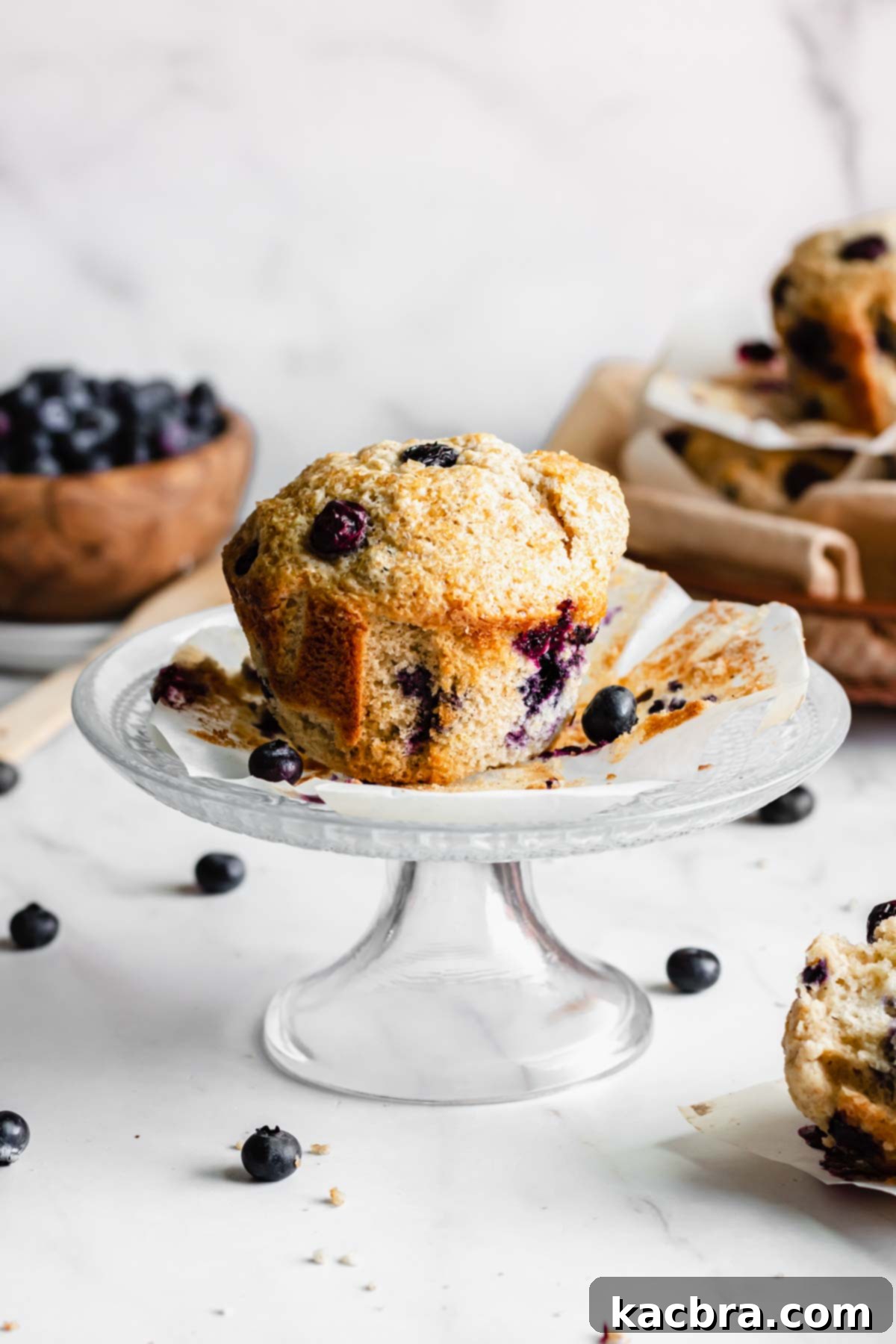 A single jumbo bakery-style blueberry muffin displayed on a charming small pedestal cake stand, highlighting its impressive size and coarse sugar topping.