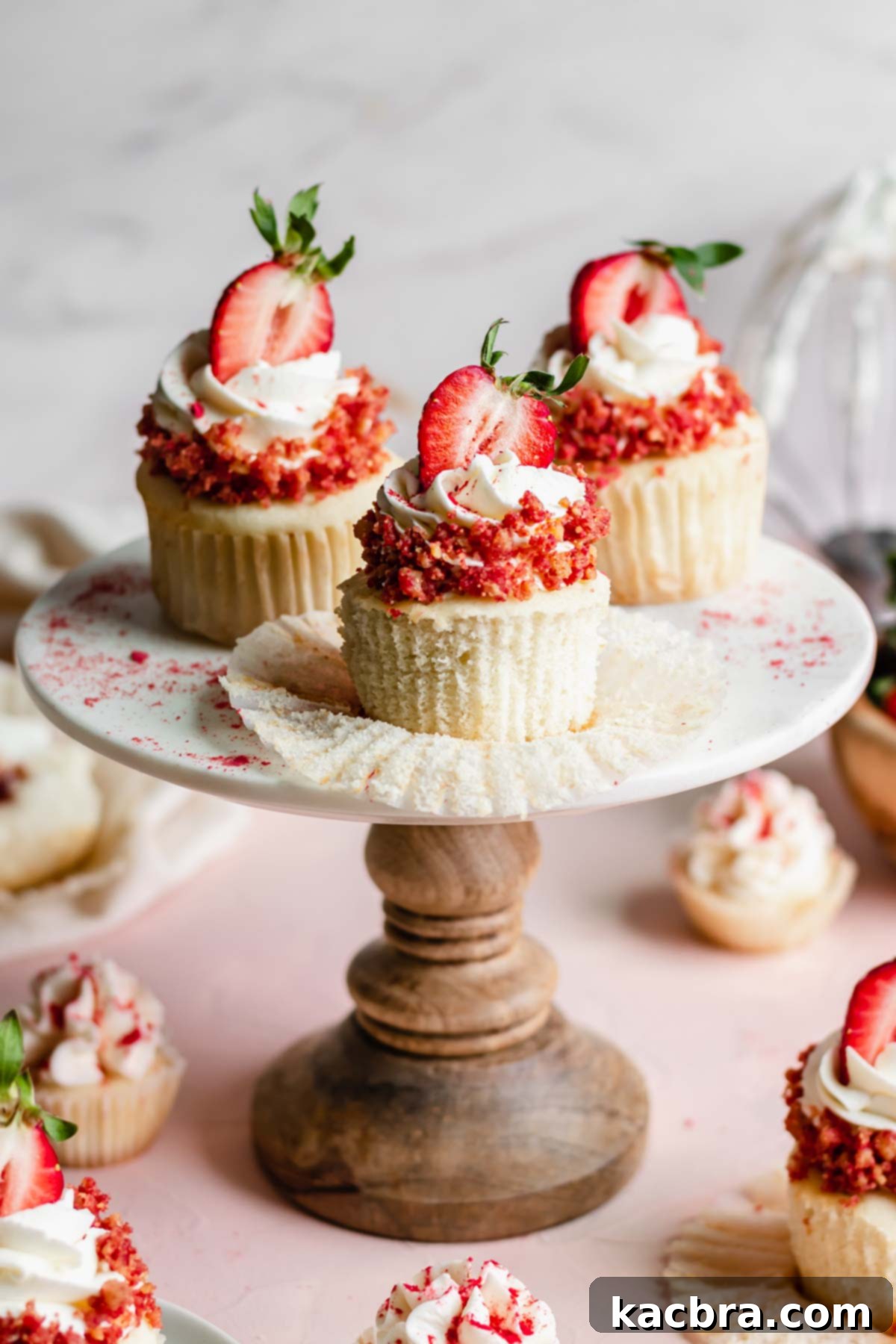 Double Strawberry Crunch Cupcakes 22 Three strawberry crunch cupcakes displayed on a cake stand, with the front cupcake revealing its removed liner to show the tender cake base beneath the frosting and crumble.