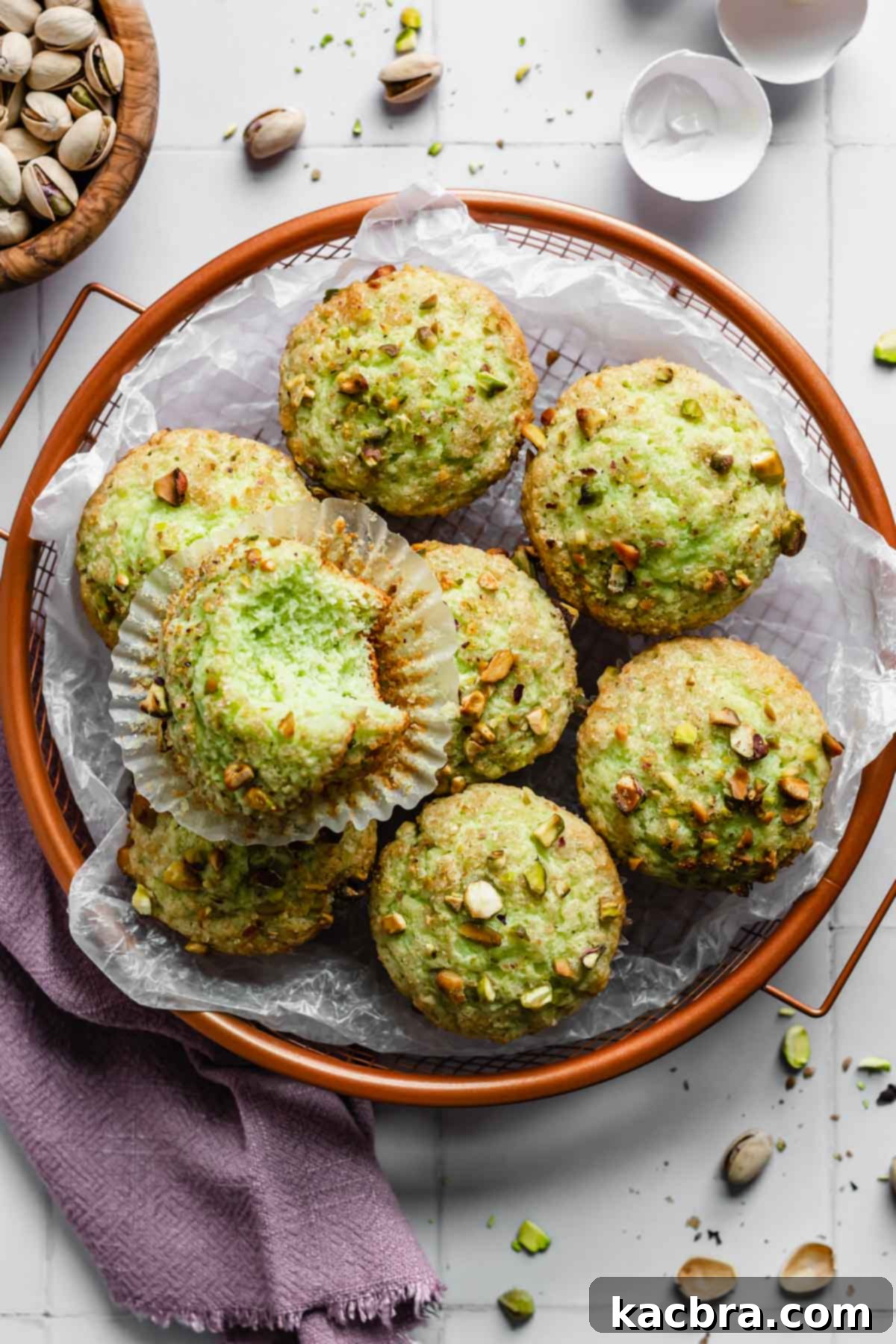 A basket overflowing with freshly baked, perfectly domed pistachio muffins, one with a bite taken out, revealing its moist, green interior.