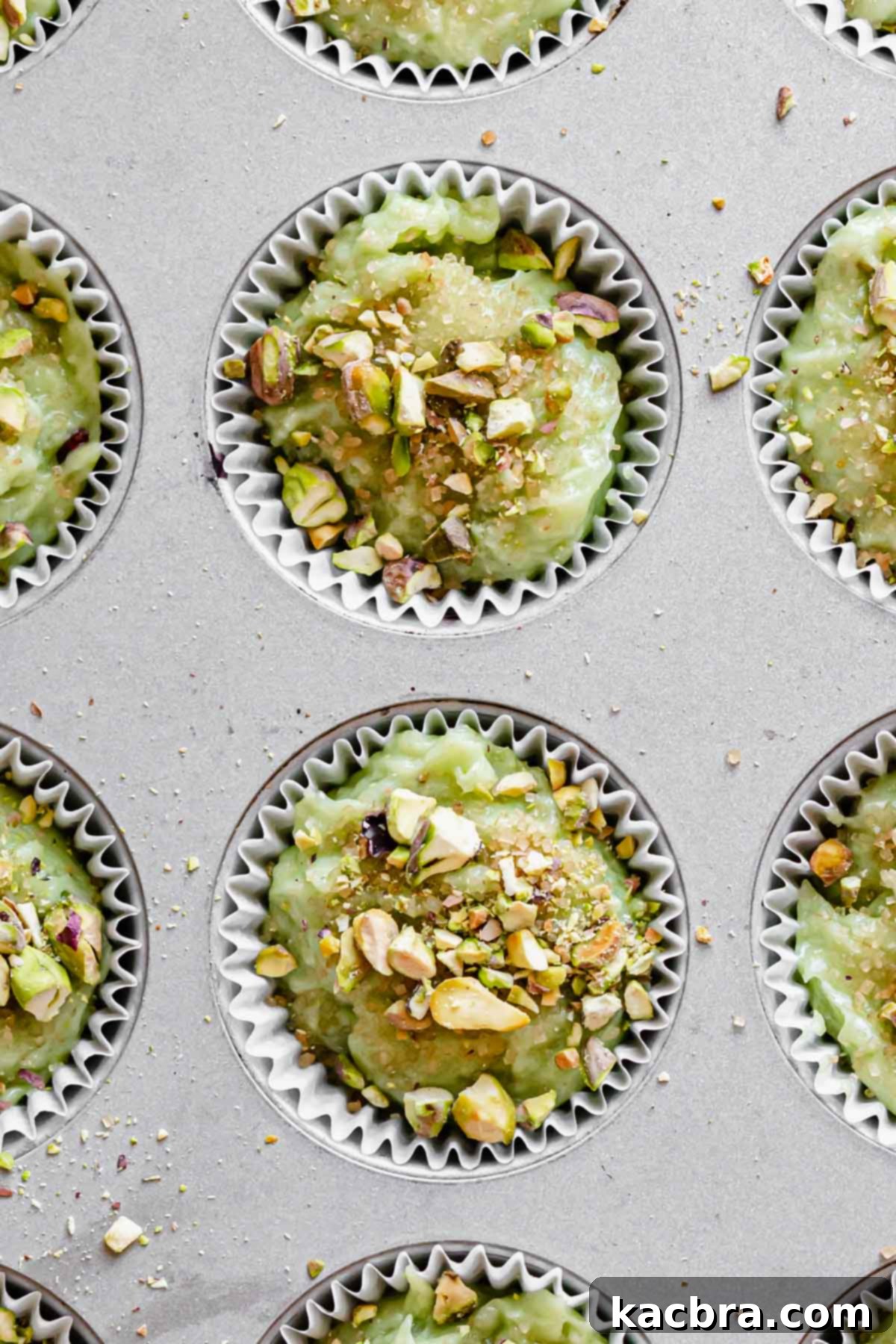 Muffin tin filled with green pistachio batter, topped with chopped pistachios and coarse sugar, ready for baking.