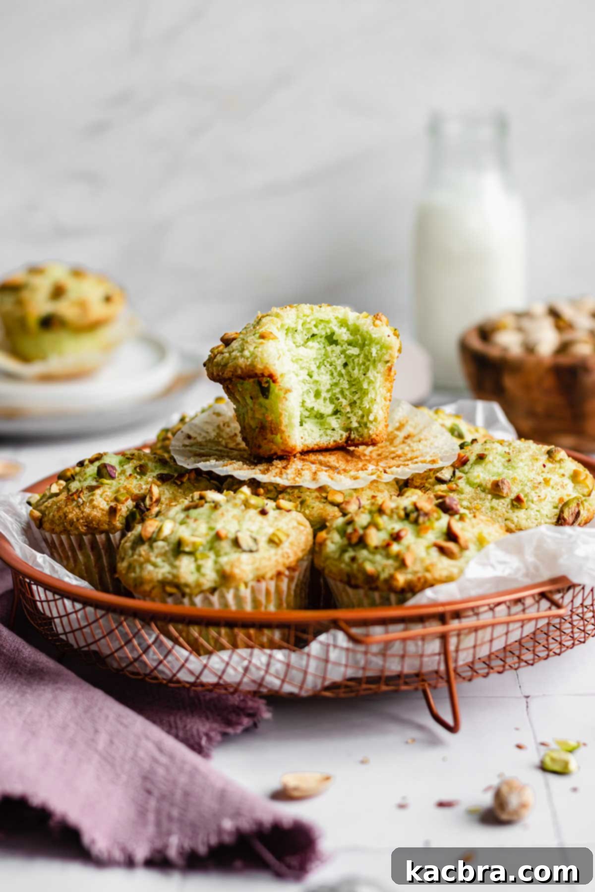 A perfectly baked pistachio muffin with a bite taken out, resting on top of a rustic basket filled with more pistachio muffins, ready to be enjoyed.