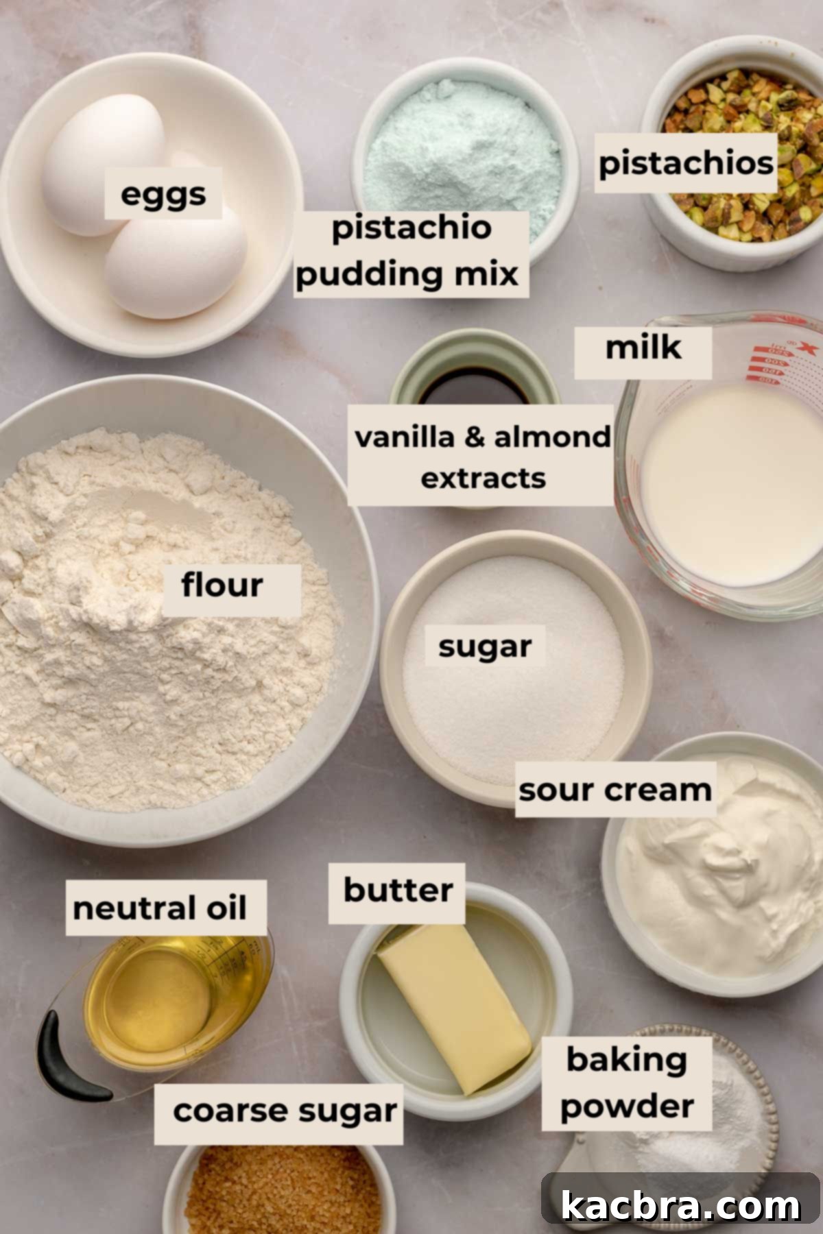 A collection of ingredients for pistachio muffins, including flour, eggs, oil, butter, sour cream, milk, pistachio pudding mix, baking powder, and pistachios arranged neatly on a surface.