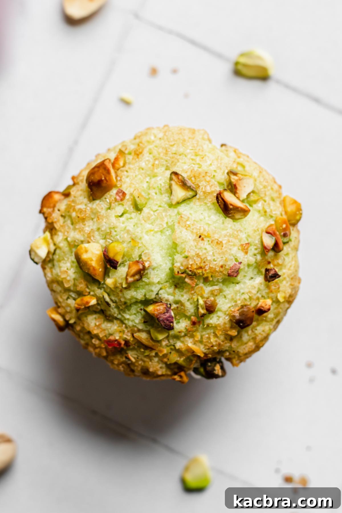 A close-up, overhead shot of a single bakery-style pistachio muffin, highlighting its perfectly domed, golden-brown top sprinkled with coarse sugar and chopped pistachios.