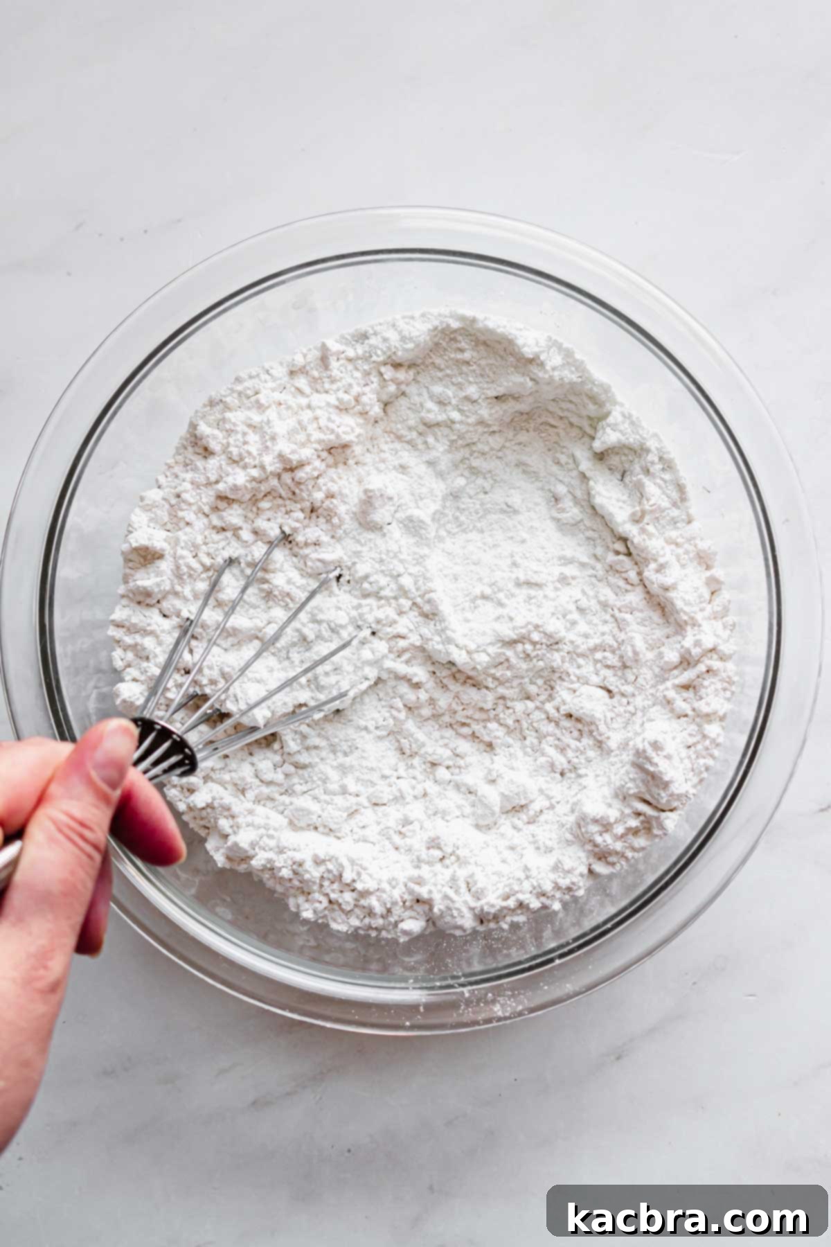 A whisk thoroughly combines dry ingredients, including flour and pistachio pudding mix, in a mixing bowl.