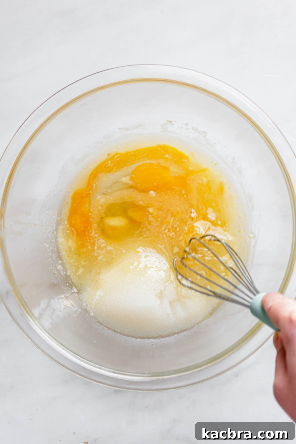 A whisk vigorously mixes the wet ingredients, including eggs, sugar, melted butter, and oil, in a large mixing bowl.