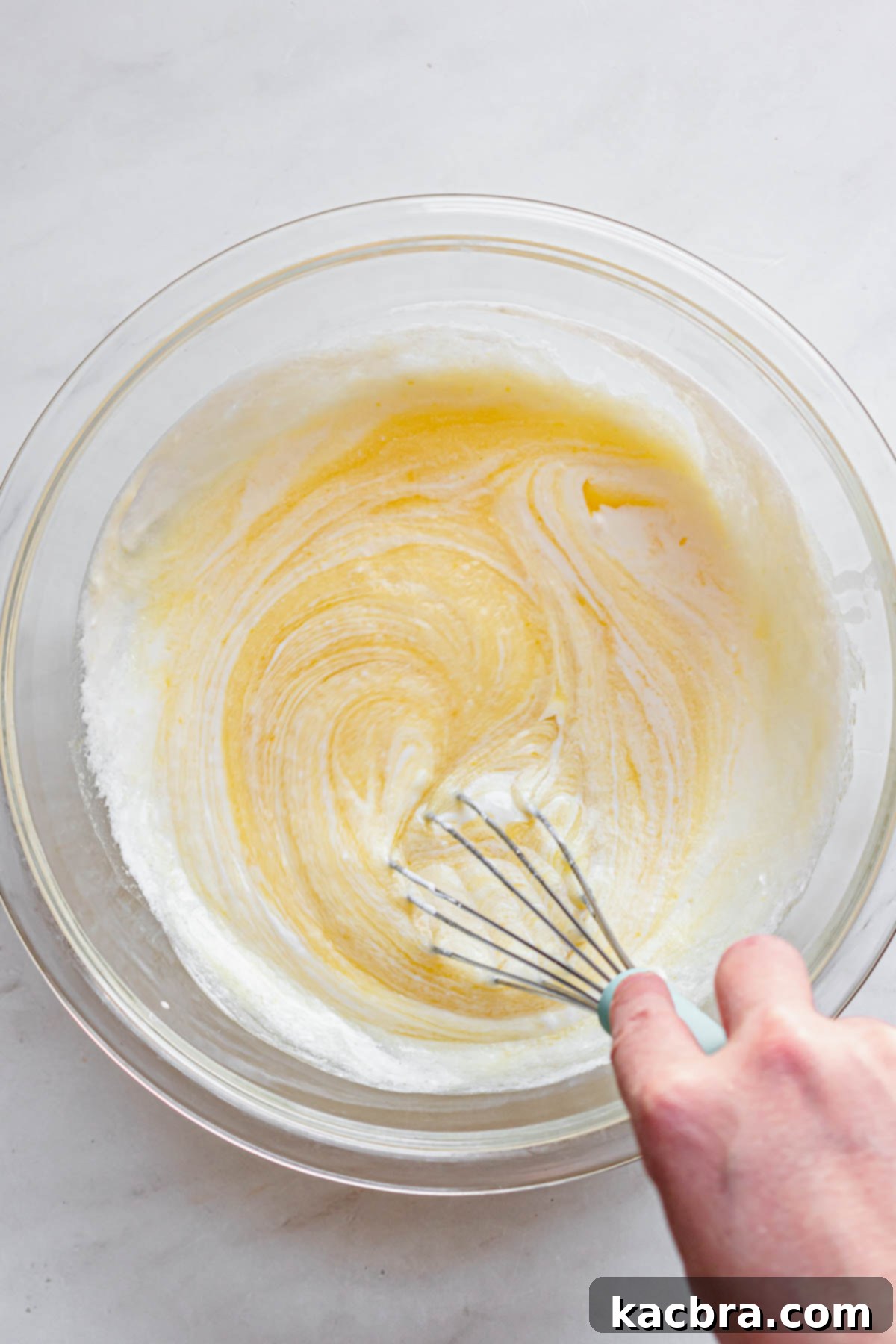 A hand holds a whisk, incorporating sour cream, whole milk, vanilla extract, and almond extract into the wet ingredients in a mixing bowl.