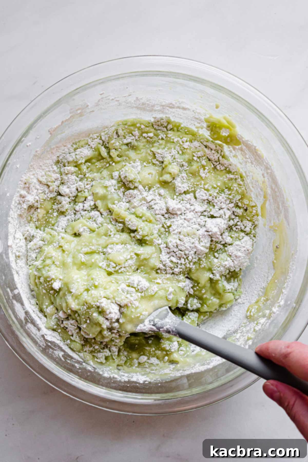 A rubber spatula carefully folds the green-hued dry ingredients into the wet ingredients in a mixing bowl, beginning to combine the batter.