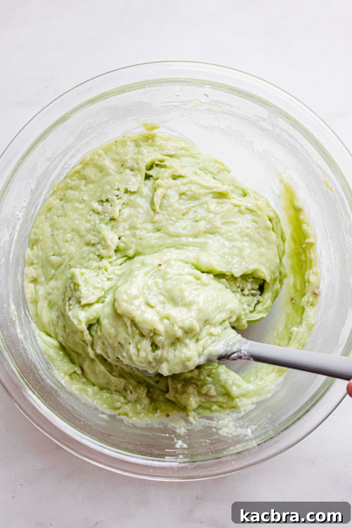 The thick, vibrant green pistachio muffin batter with a rubber spatula lifting a portion to show its consistency.