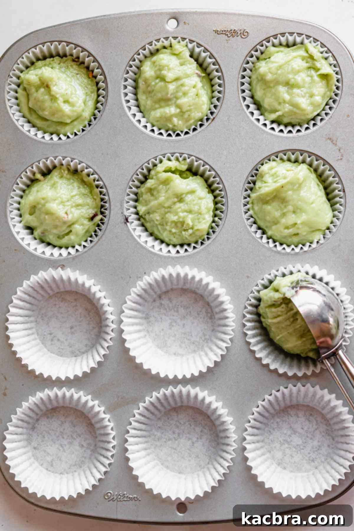 A cookie scoop fills muffin liners in a muffin tin with the thick green pistachio batter, filling each cup to the brim.