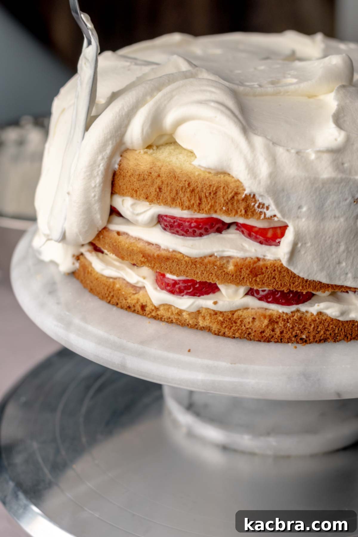 A spatula spreading whipped cream all over the assembled layer cake.