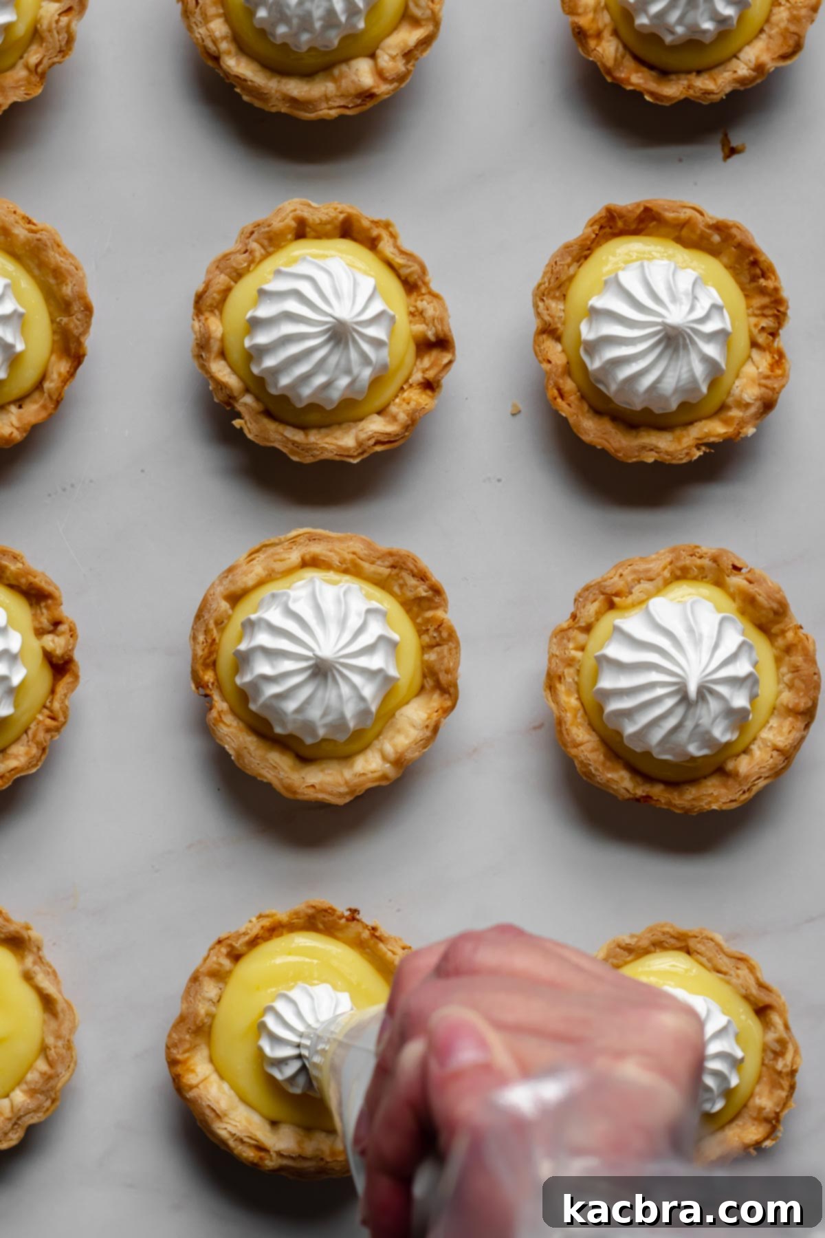 Zingy Lemon Meringue Kisses 8 Hands piping elegant swirls of Swiss meringue onto a mini lemon tart.