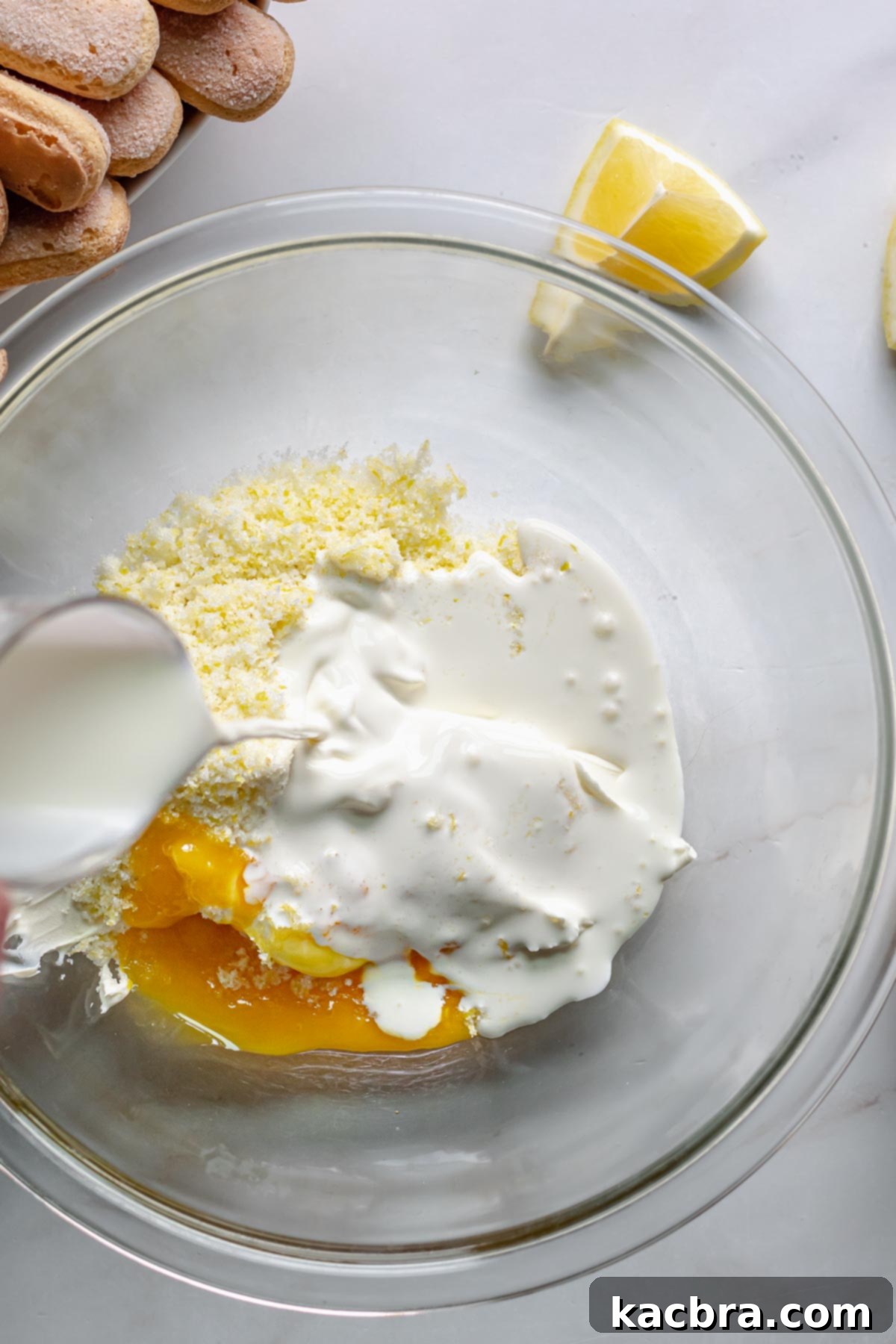 Heavy cream being added to mascarpone, lemon sugar, and egg yolks in a mixing bowl.