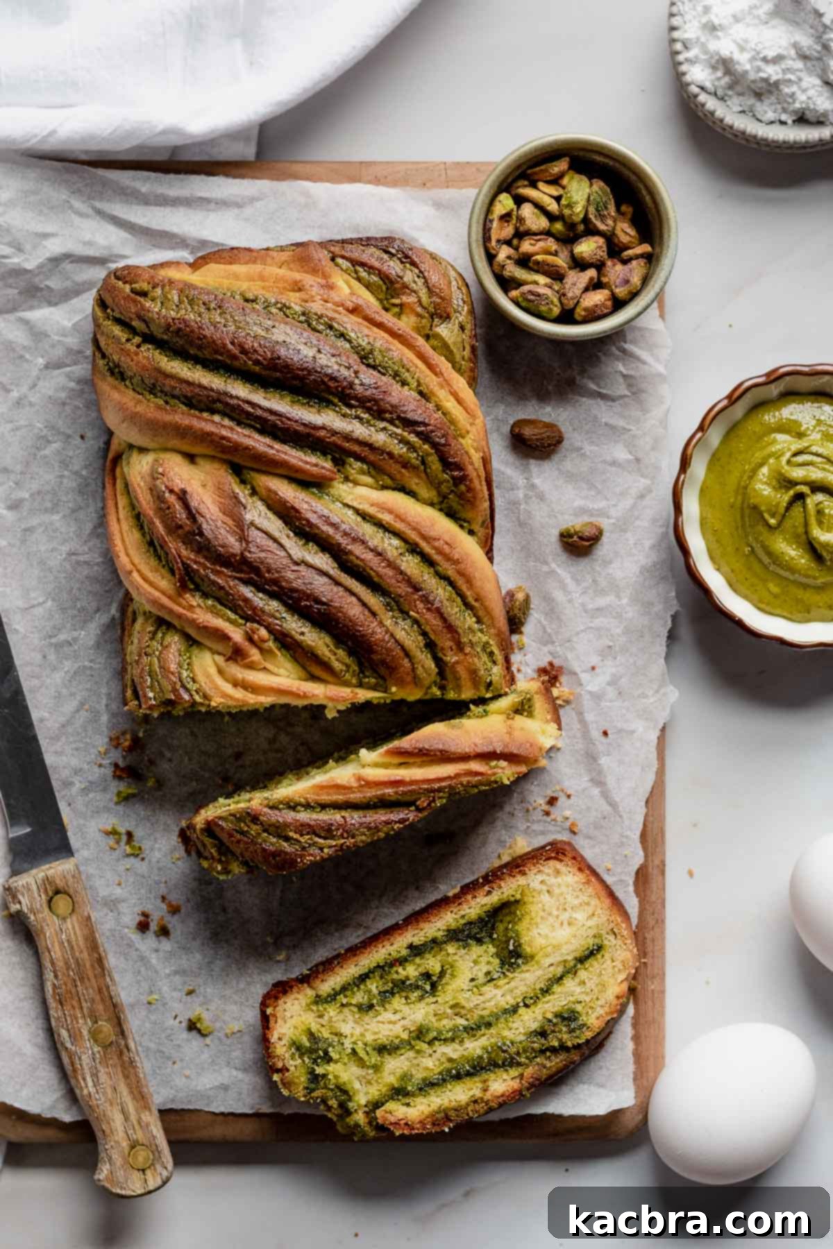 A freshly baked Pistachio Babka loaf, beautifully braided and golden brown, with two inviting slices cut from it, revealing the vibrant green swirls of pistachio filling within. The loaf sits on a rustic wooden surface, hinting at homemade goodness.