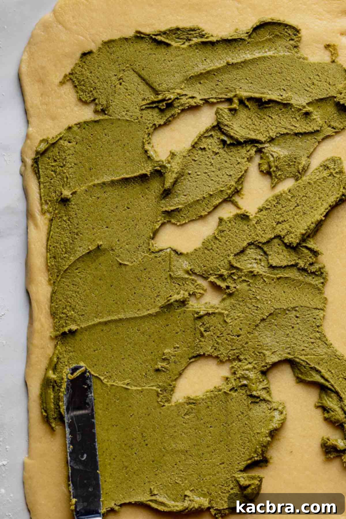 A spatula delicately spreading the vibrant pistachio filling across the flattened babka dough in an even layer.